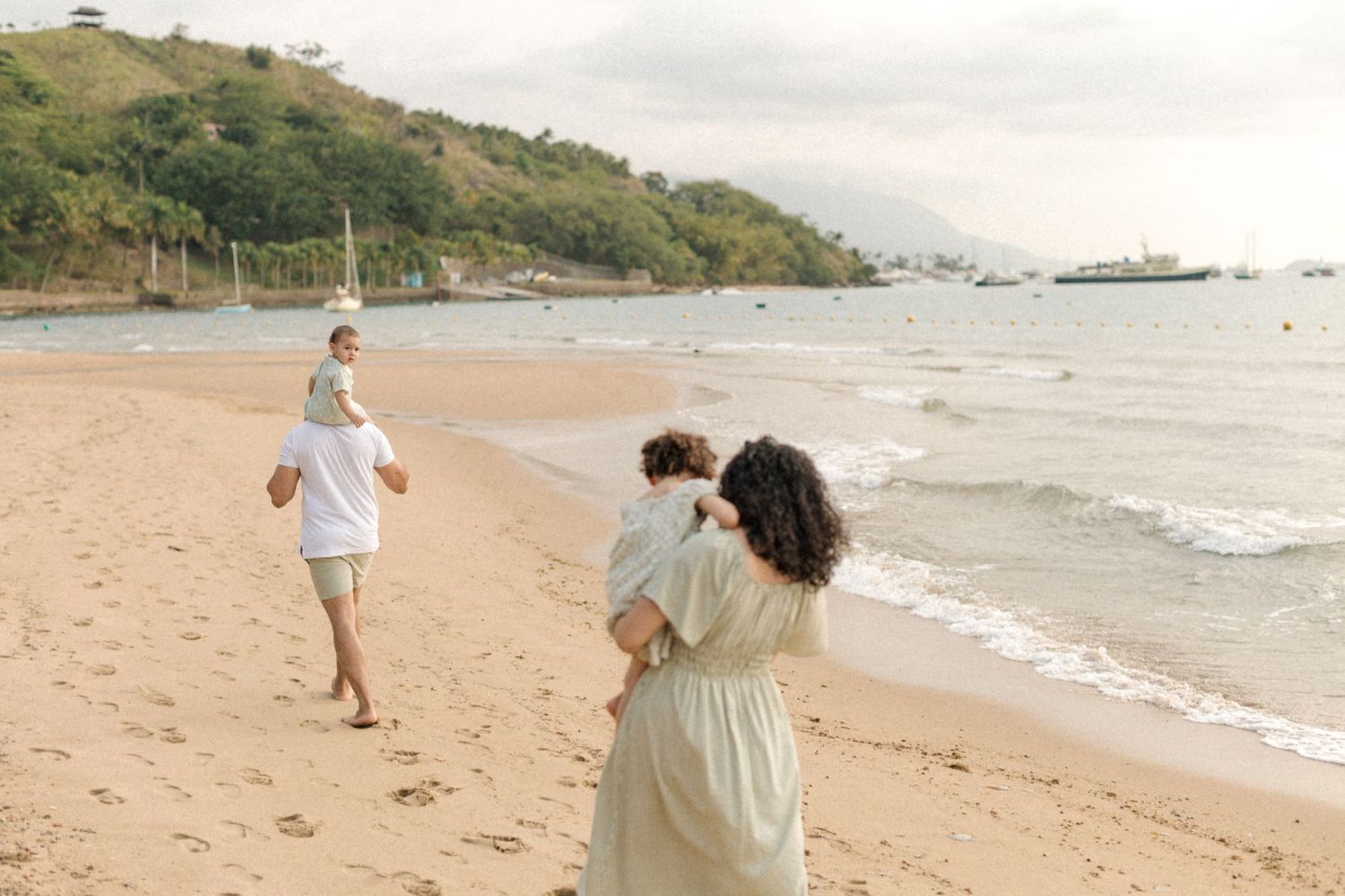 People enjoying a sunny day at a sandy beach with hills and boats visible in the distance.