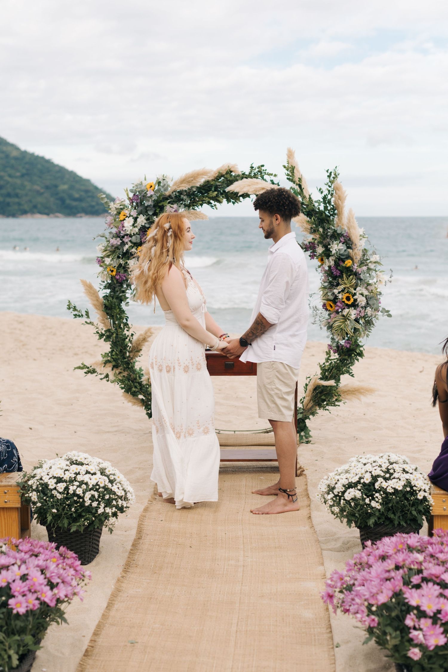 Couple stands under floral arch during beach wedding with ocean and mountains in background.