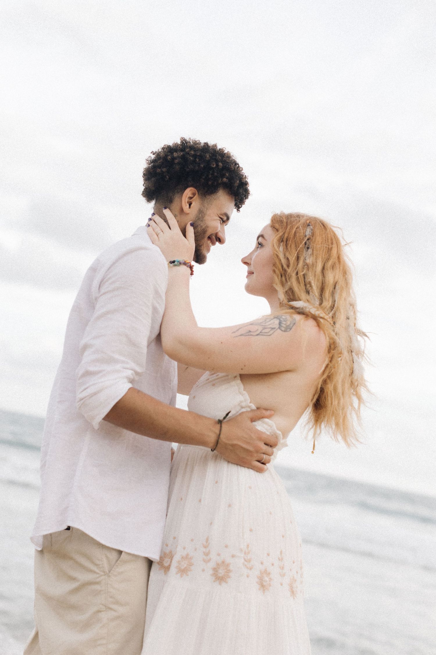 A loving couple shares an intimate moment on the beach while wearing coordinated white and beige outfits.