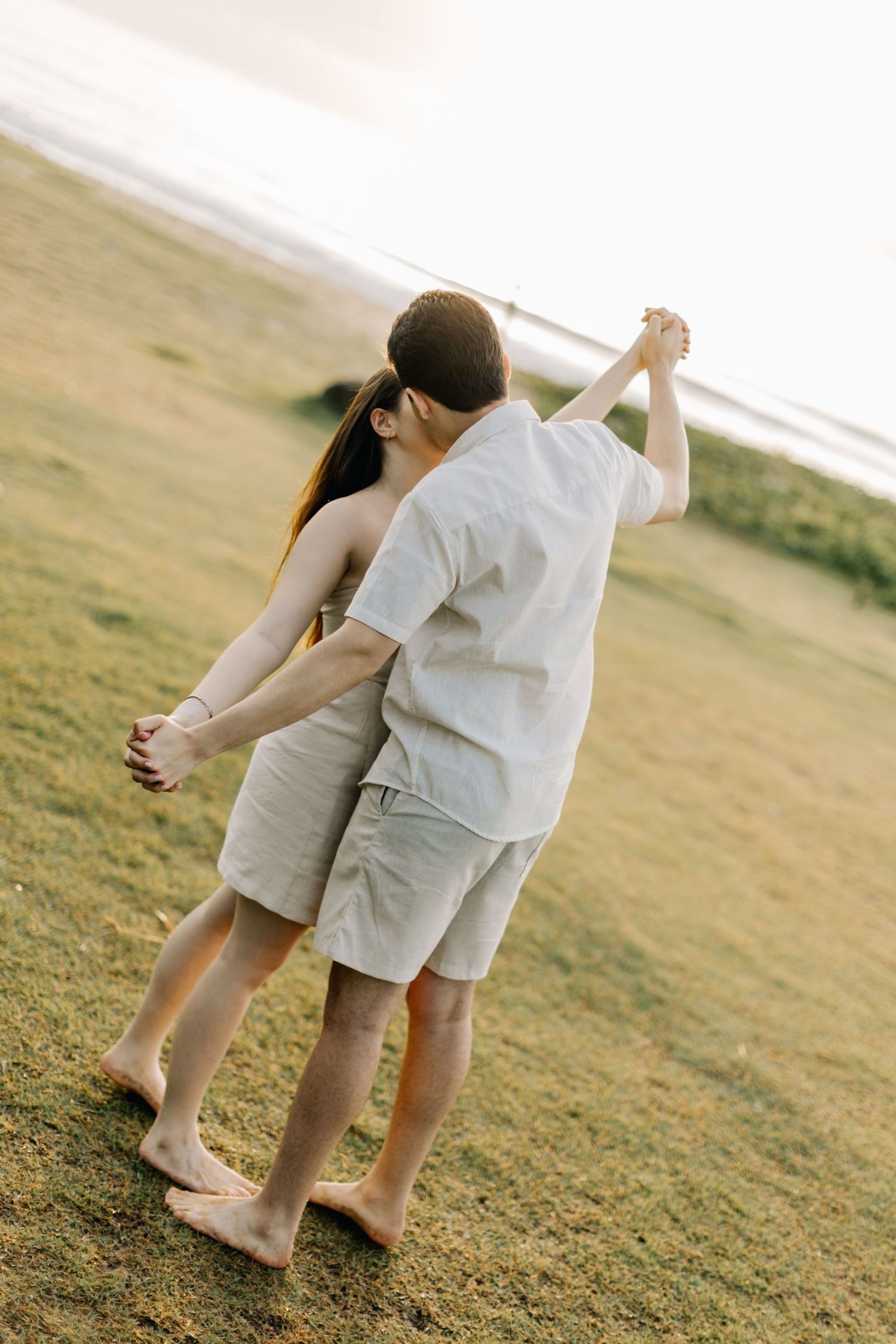 Dancing couple in light colored outfits sharing romantic moment on grassy hillside.