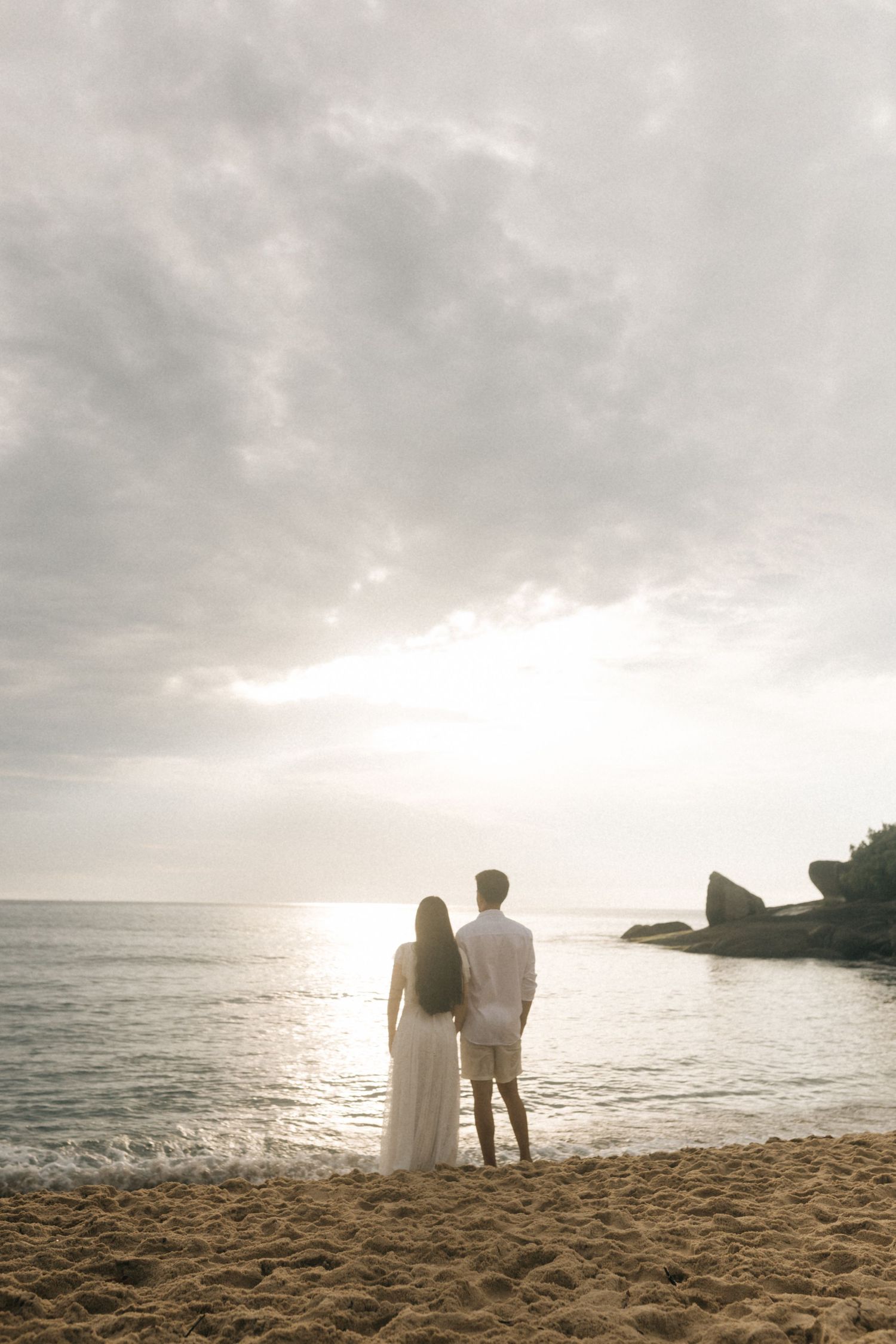 A couple stands silhouetted against a dramatic sunset on a sandy beach with cloudy skies and rocky coastline.