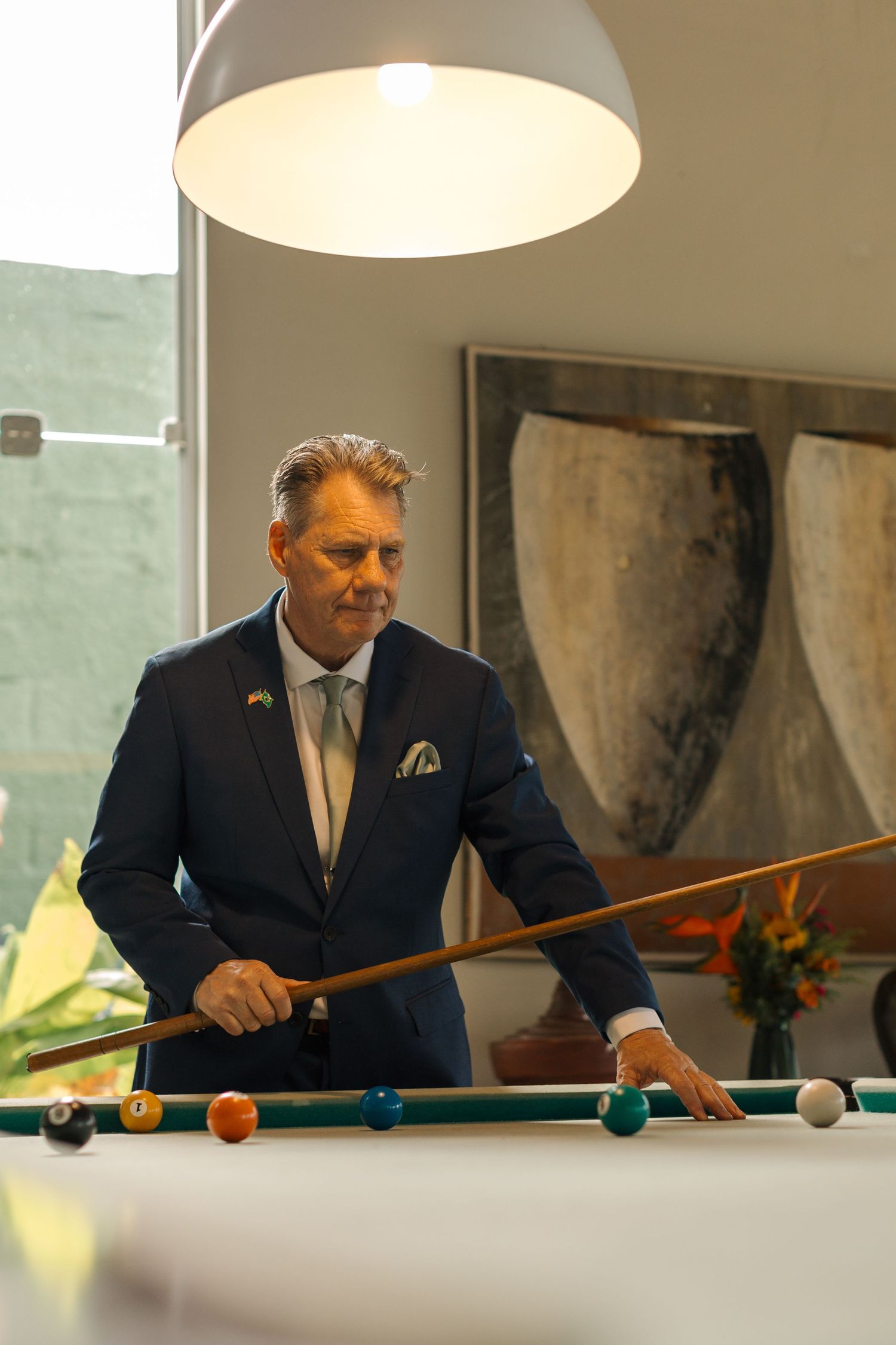 A person in a navy suit plays billiards at an elegant table under pendant lighting.