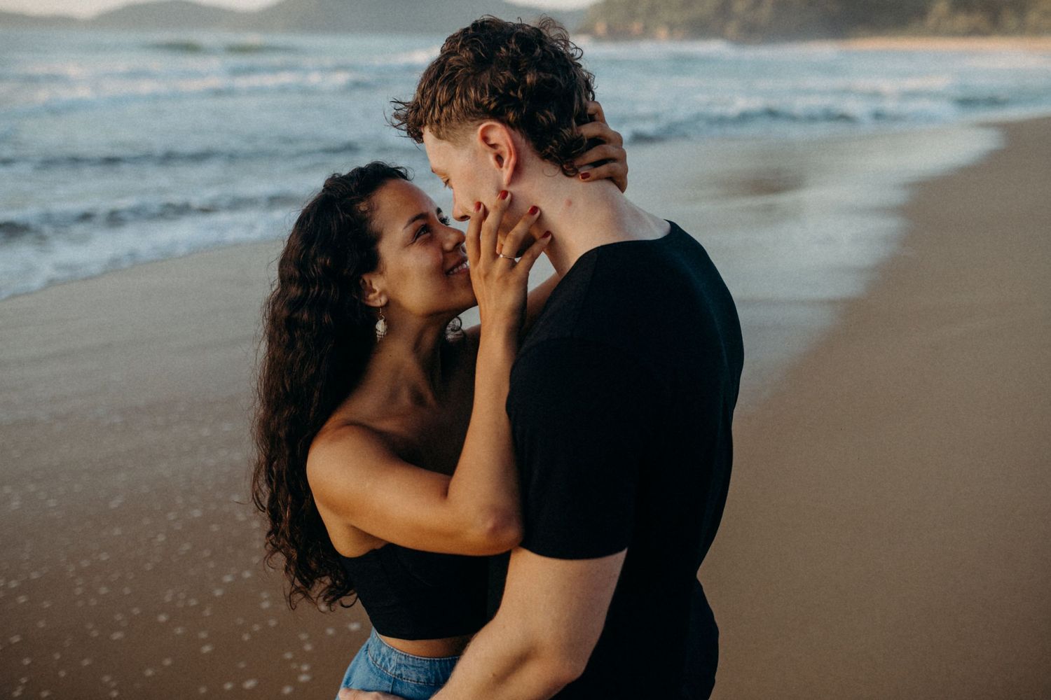 Intimate couple embraces on sandy beach with ocean waves and cliffs in background.