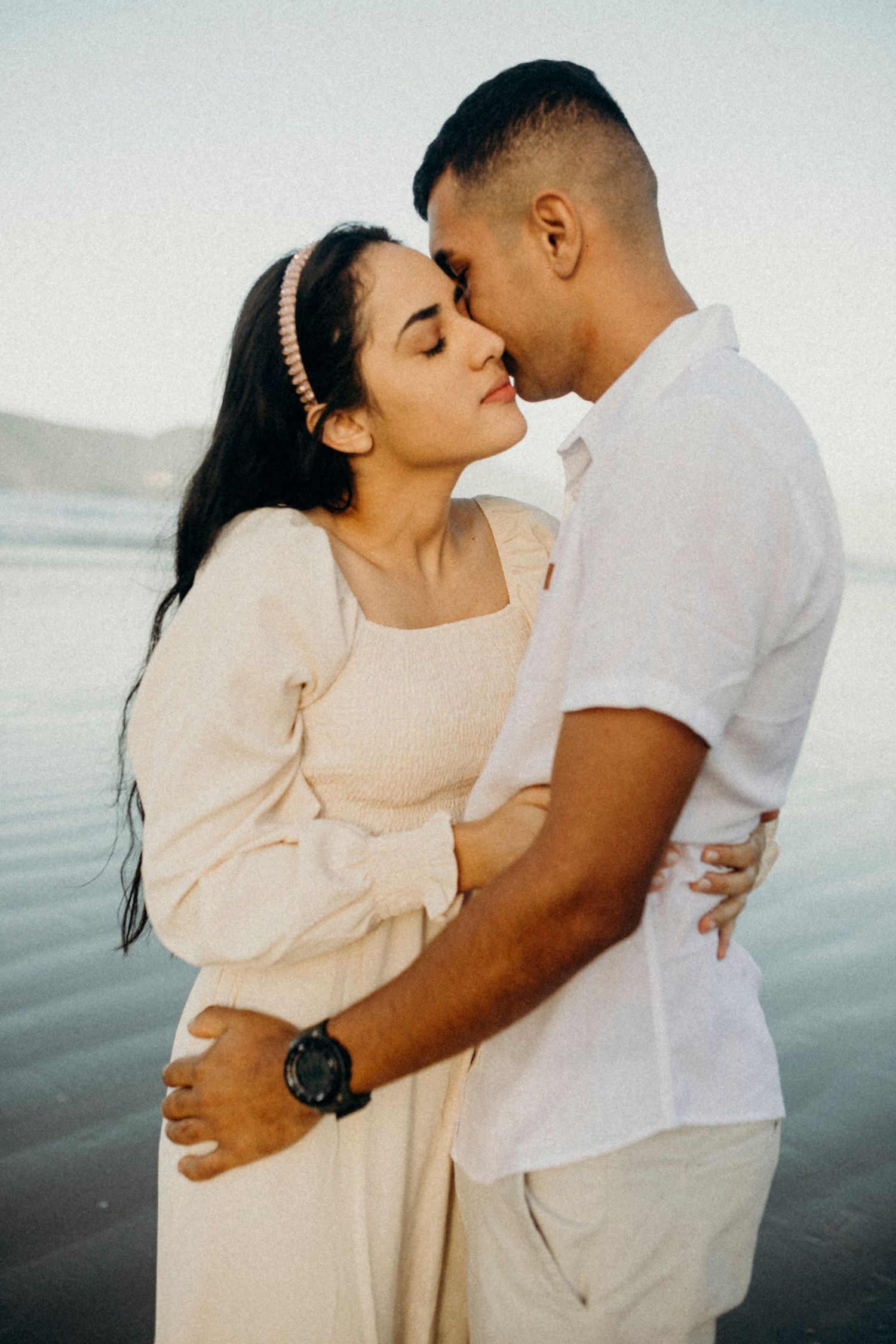 A romantic beachside portrait of a couple in white clothing embracing.