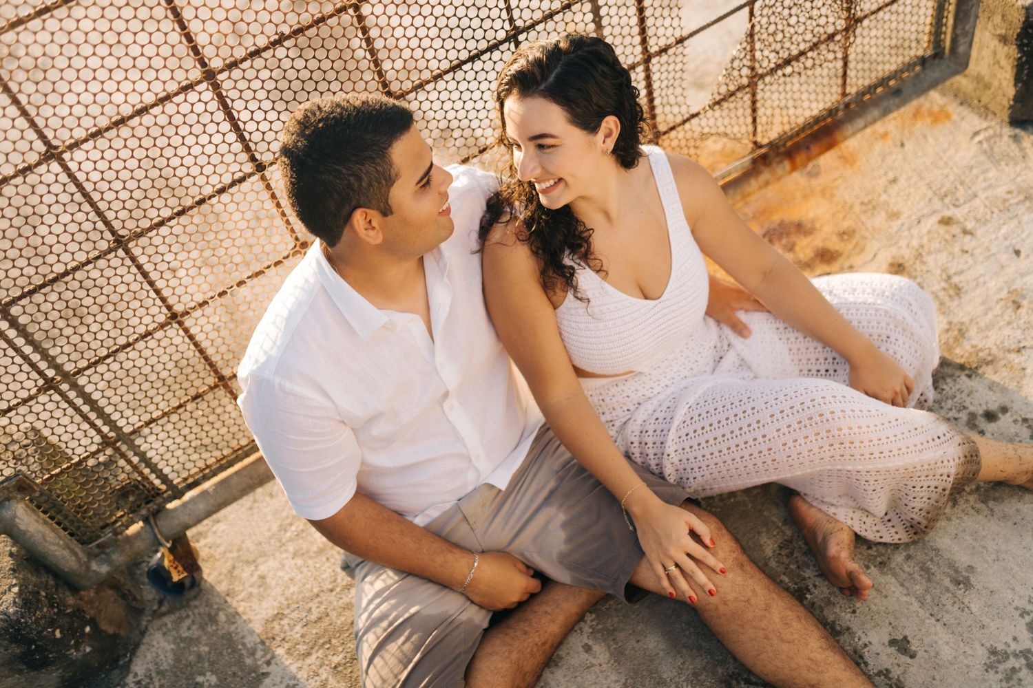 Couple in white outfits sharing an intimate moment while sitting against a textured wall in warm lighting.