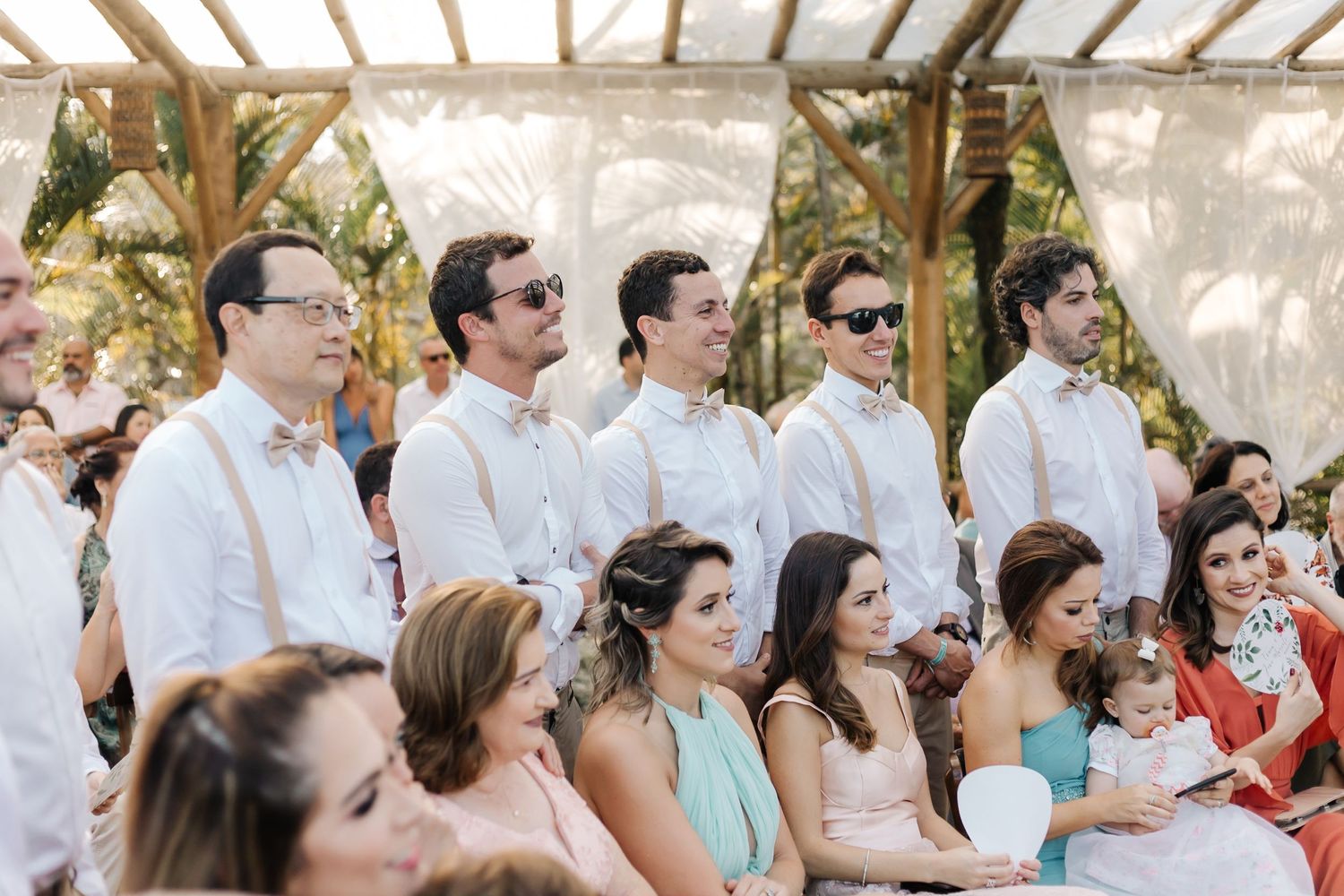 Wedding guests seated outdoors under a wooden pergola structure during a ceremony.