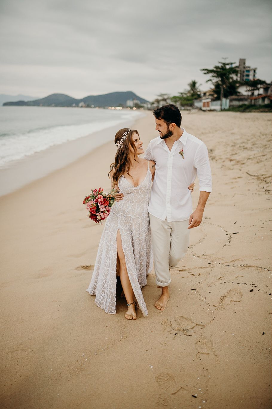 Newlyweds walking barefoot on the beach with mountains in the background.