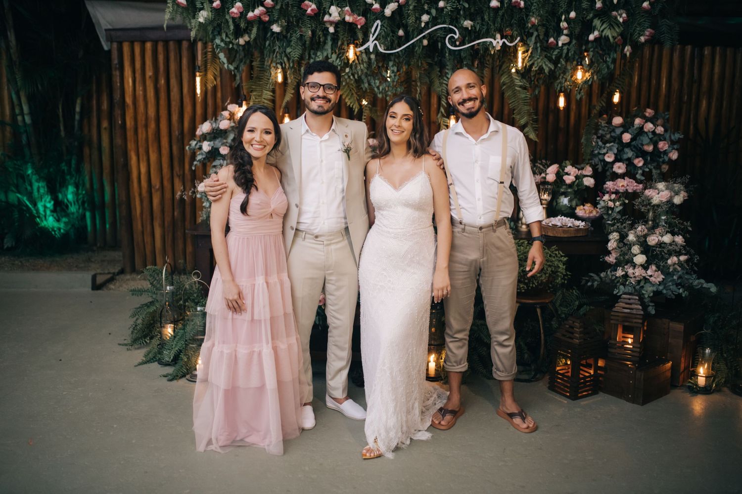 Wedding party in pastel pink and beige attire pose together against flower wall with lanterns.