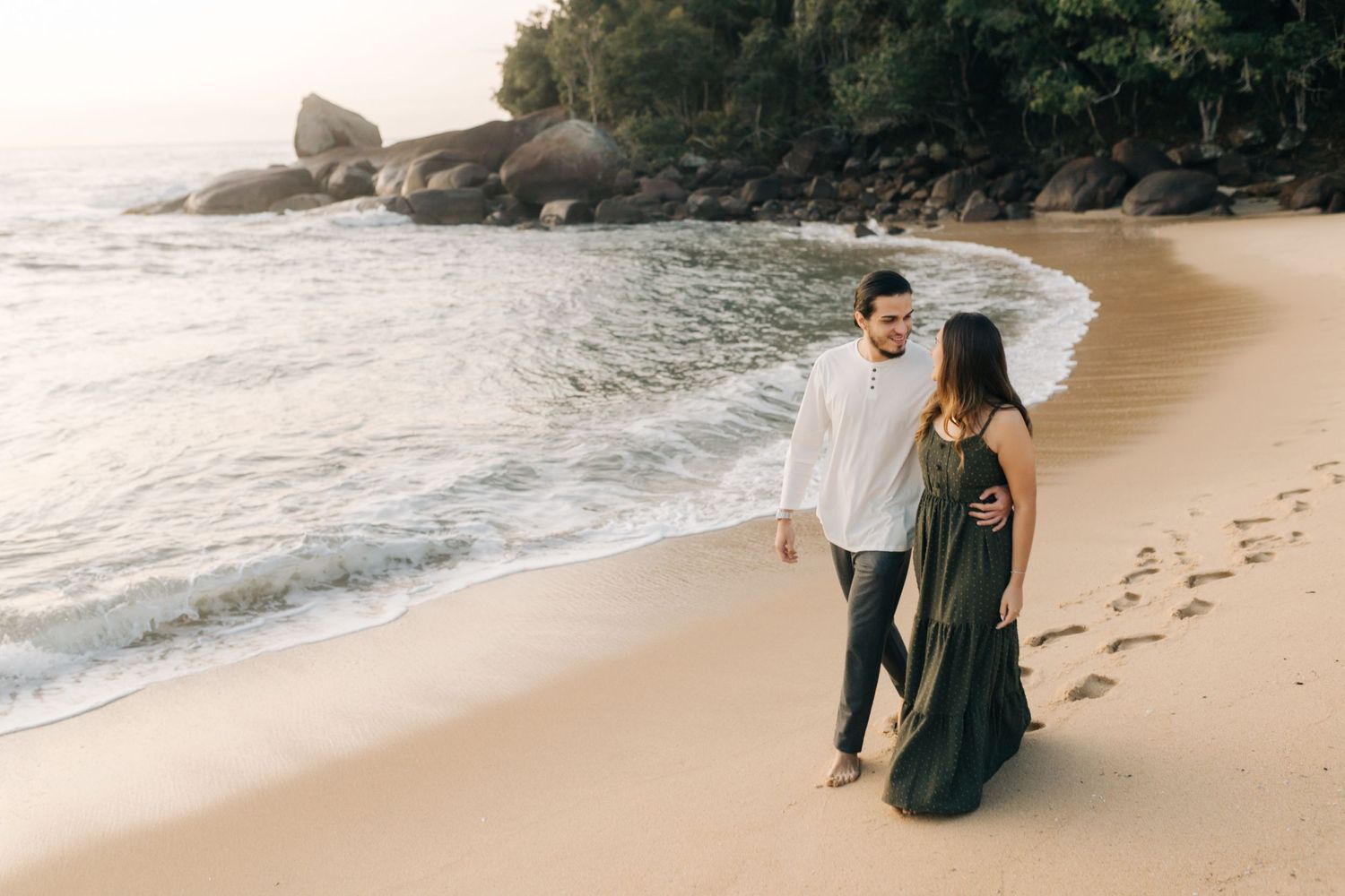Couple walking along beach leaving footprints in the sand with rocky coastline in background.