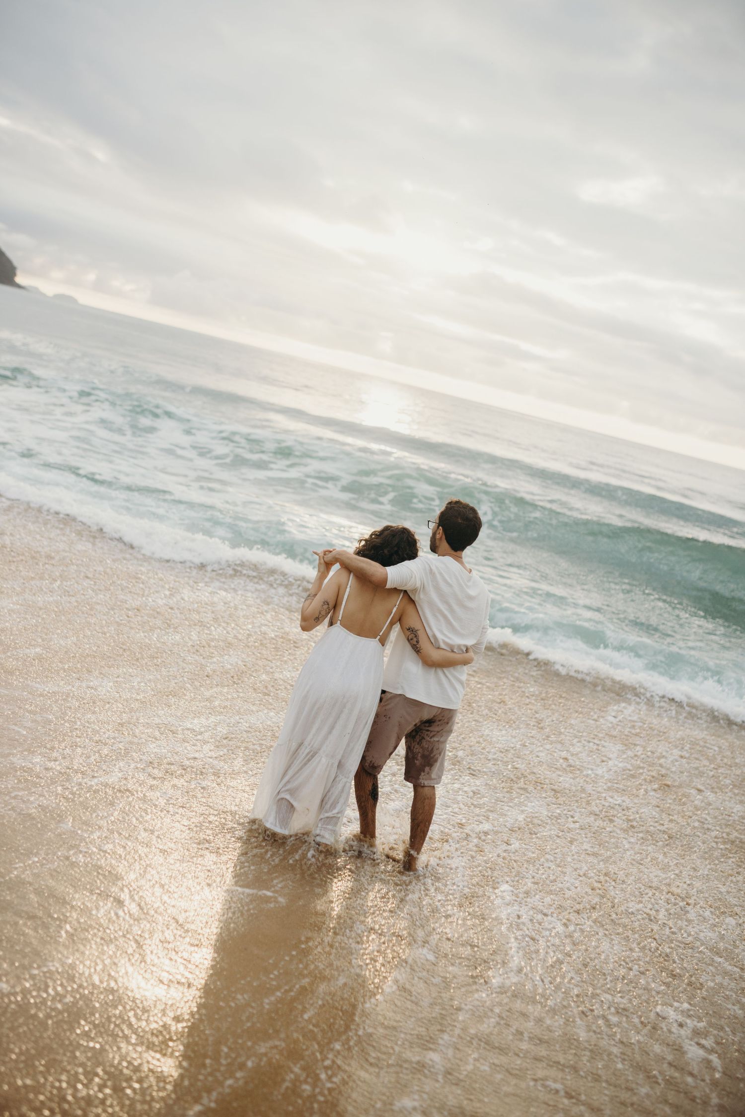 A couple stands together watching the waves roll in at sunset.