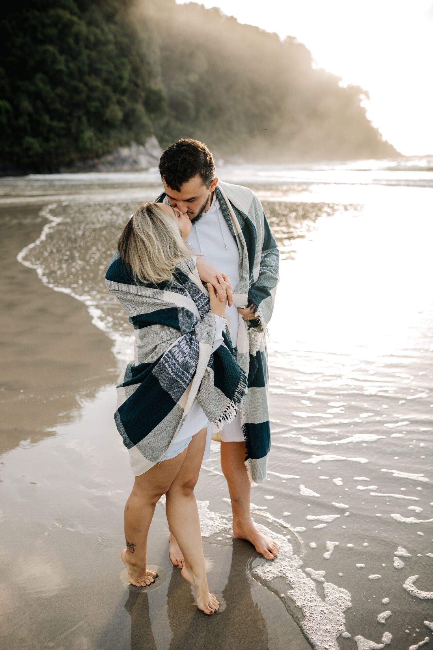 A couple shares romantic moments on a misty beach shoreline during golden hour.