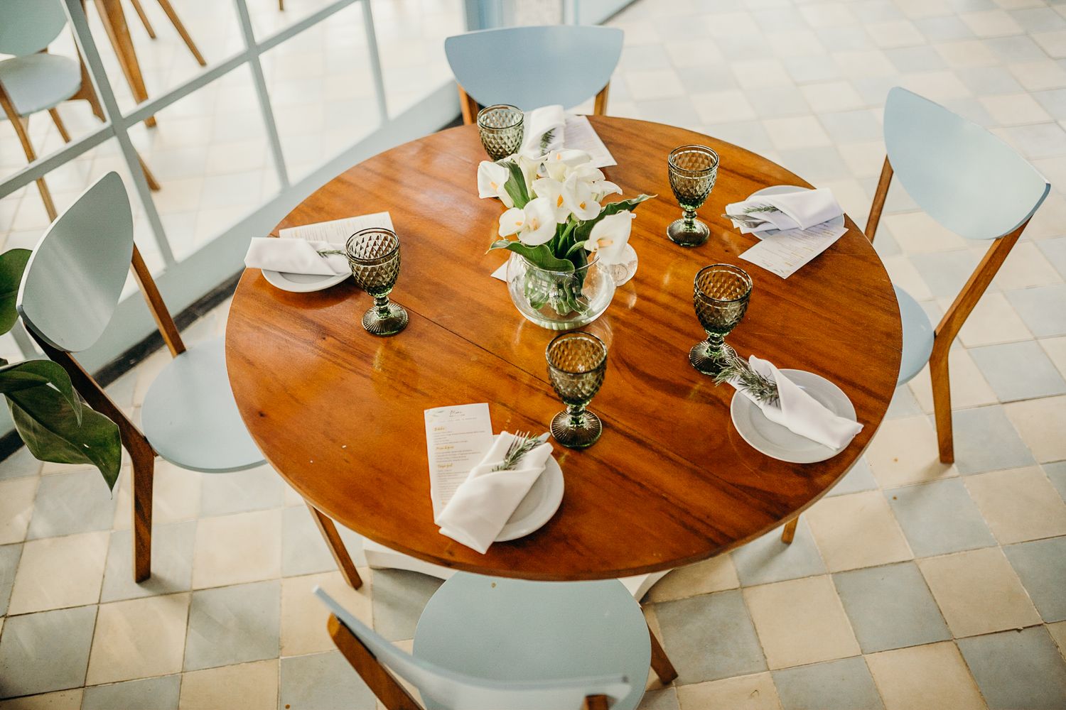 A round wooden dining table set with plates and glasses, surrounded by light blue chairs.