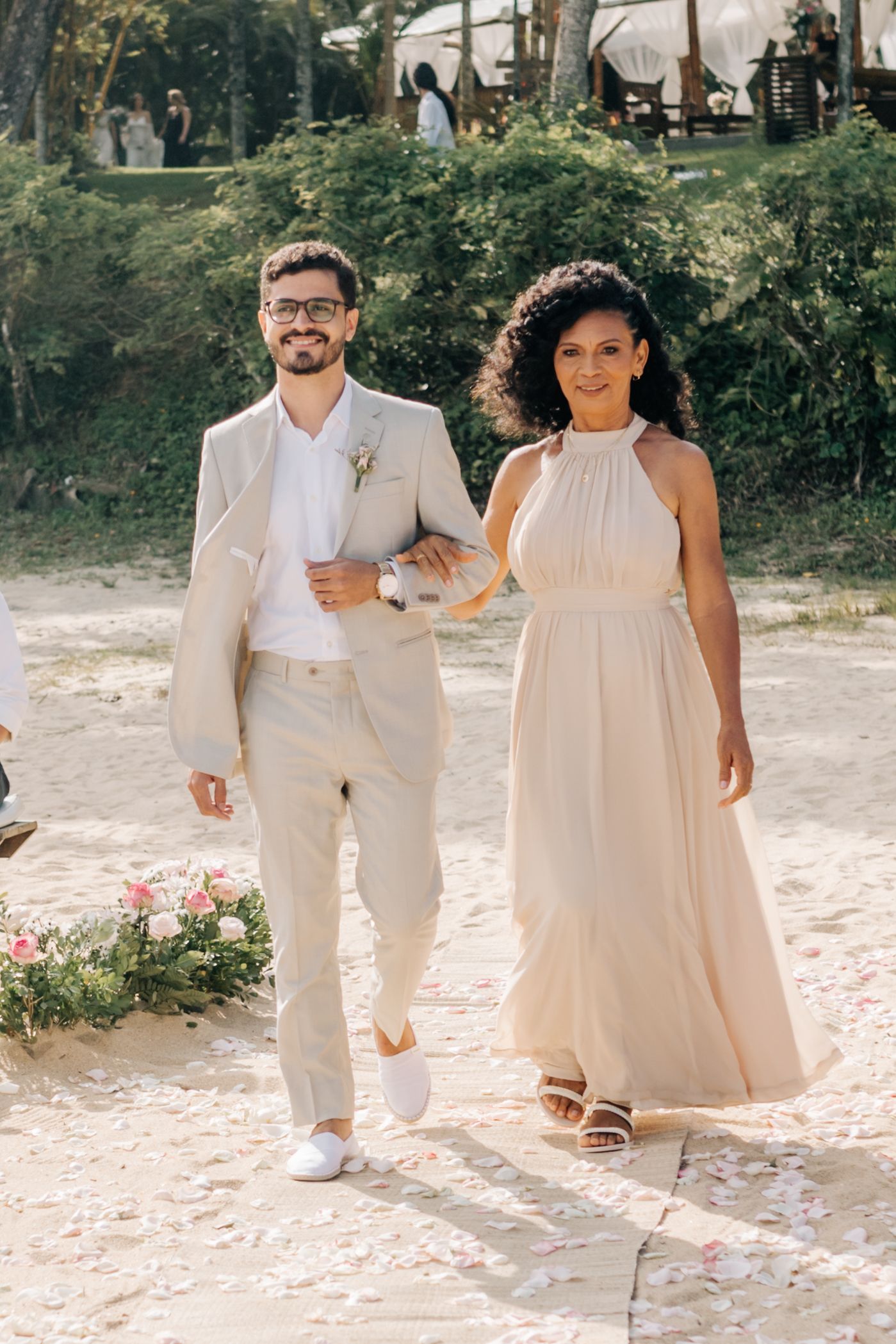A couple poses outdoors wearing light-colored wedding attire, walking arm in arm.