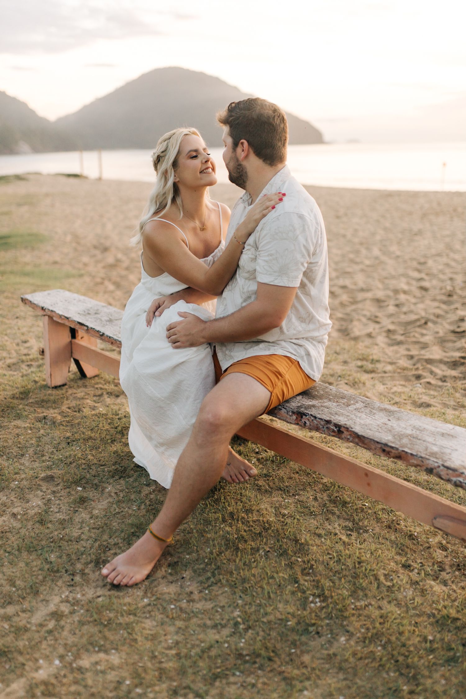 A couple shares a romantic moment on a beach bench with mountains visible in the background.