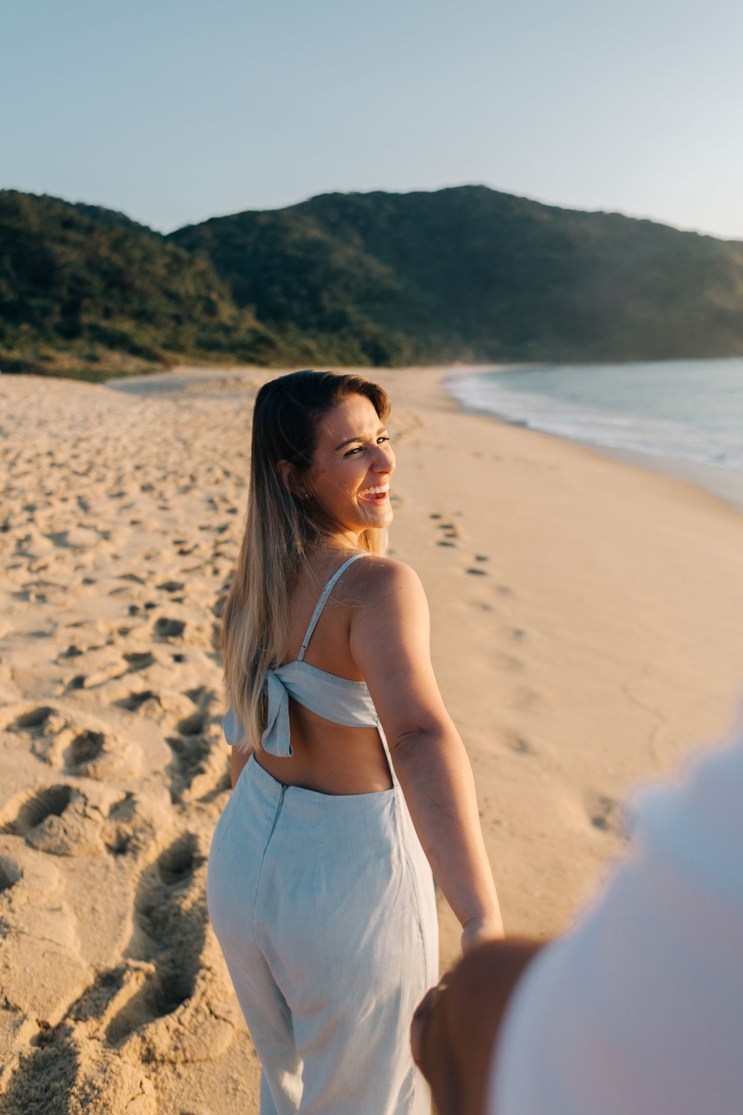 Woman in white two-piece outfit poses on beach with mountains and ocean in background.