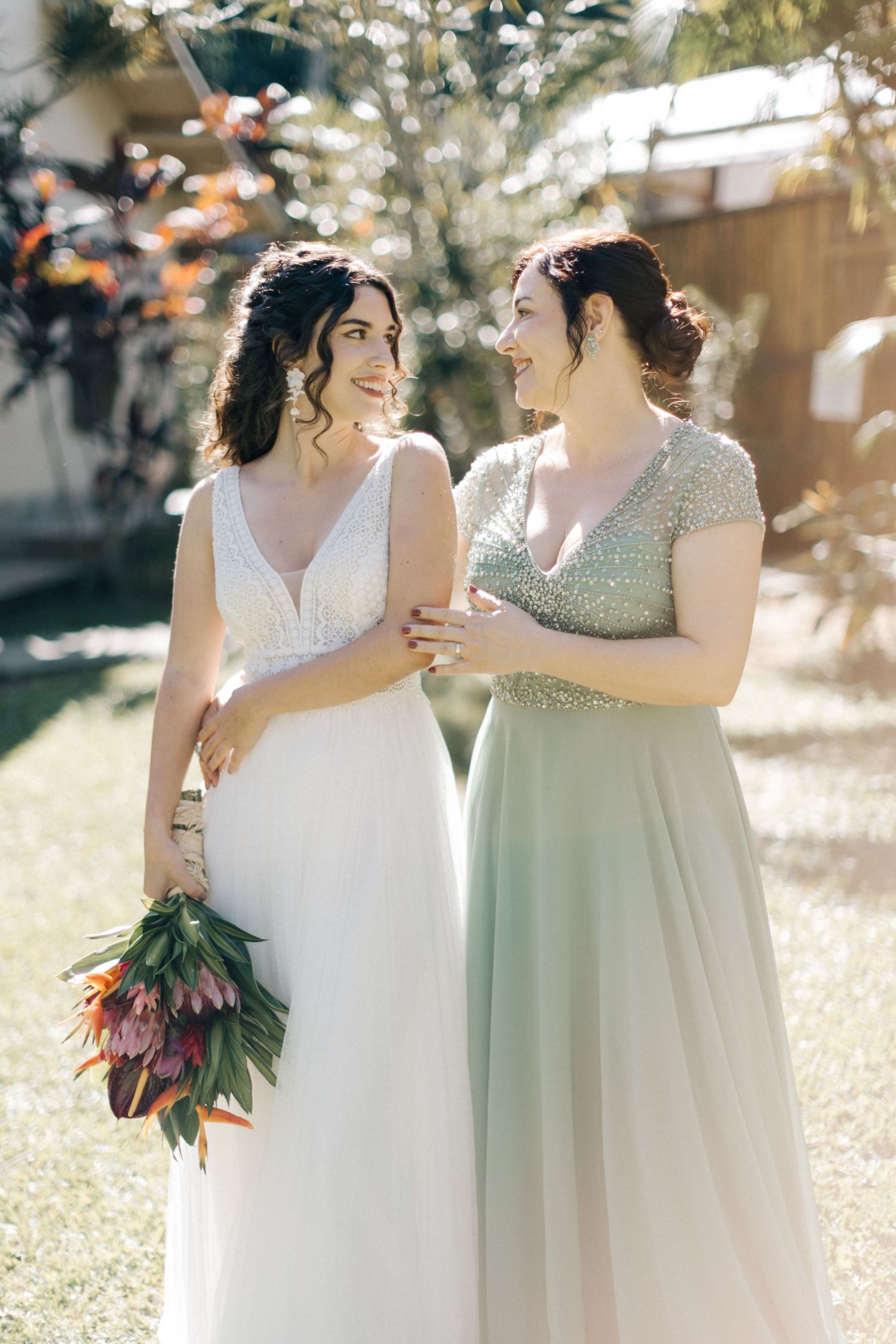 Two people in wedding attire share a joyful moment while holding a colorful bouquet in an outdoor garden.