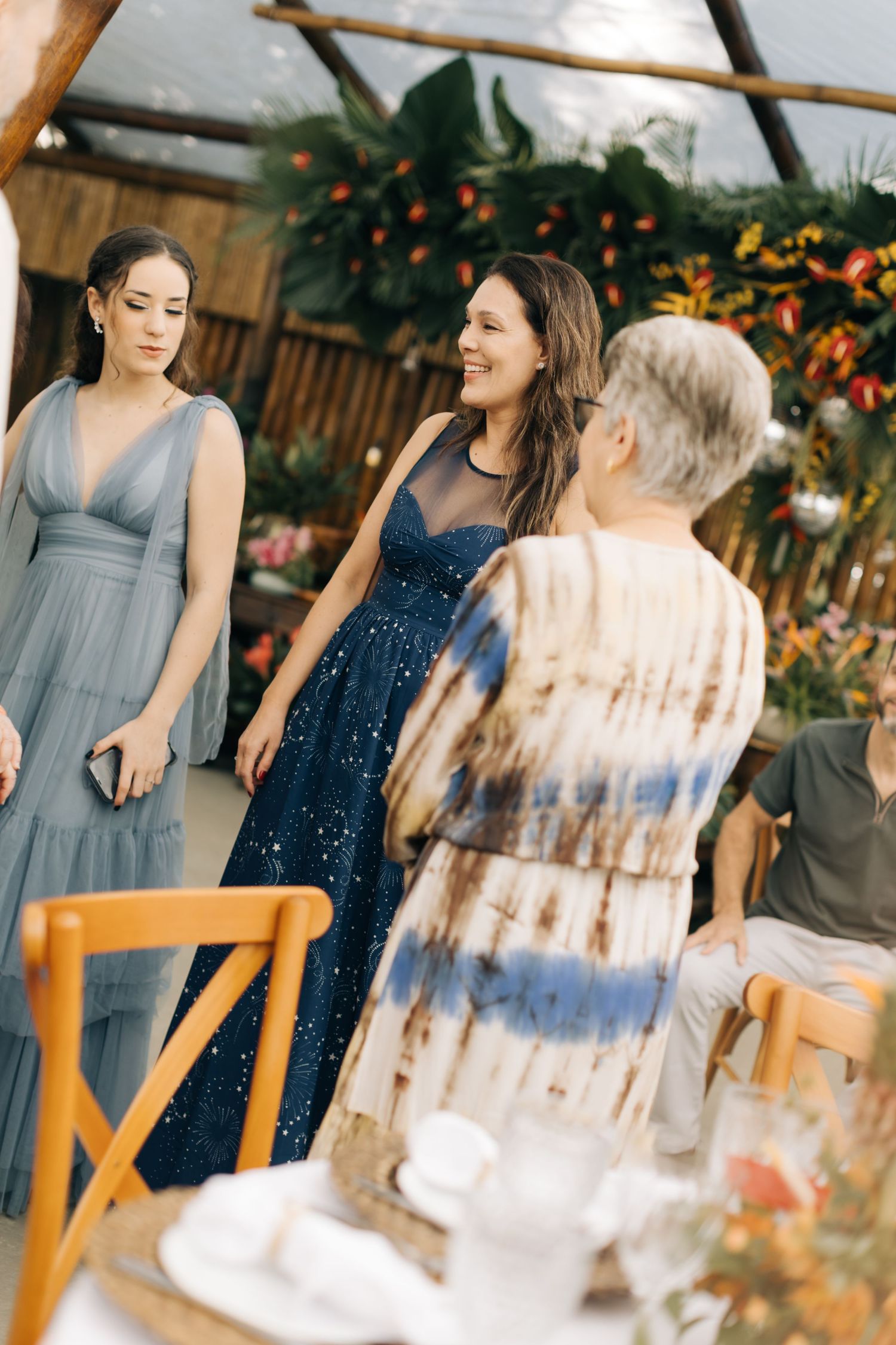 Three women in elegant dresses gather at an outdoor event with floral decorations in the background.