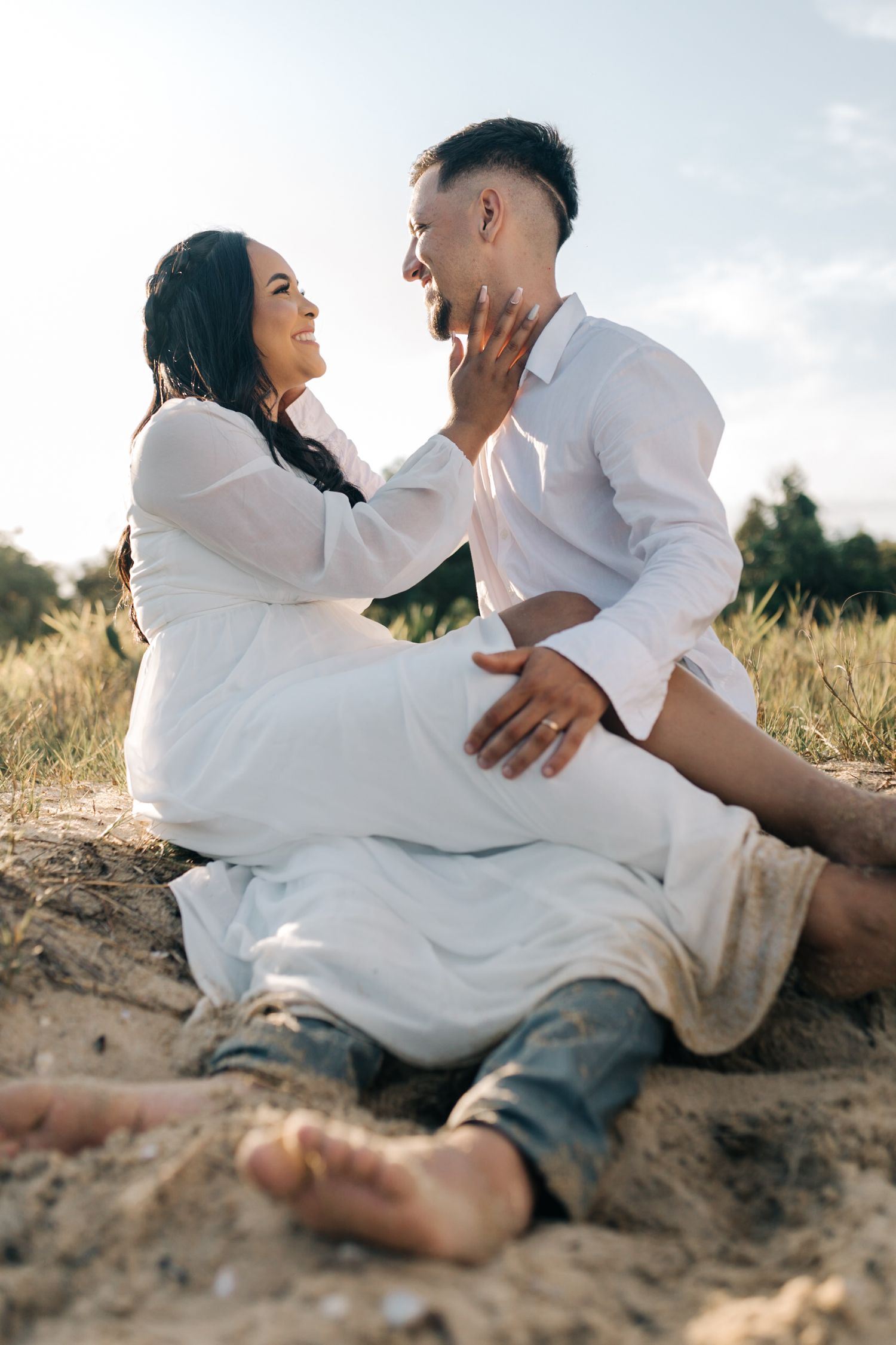 Couple shares tender moments on sandy beach while dressed in coordinating white outfits.