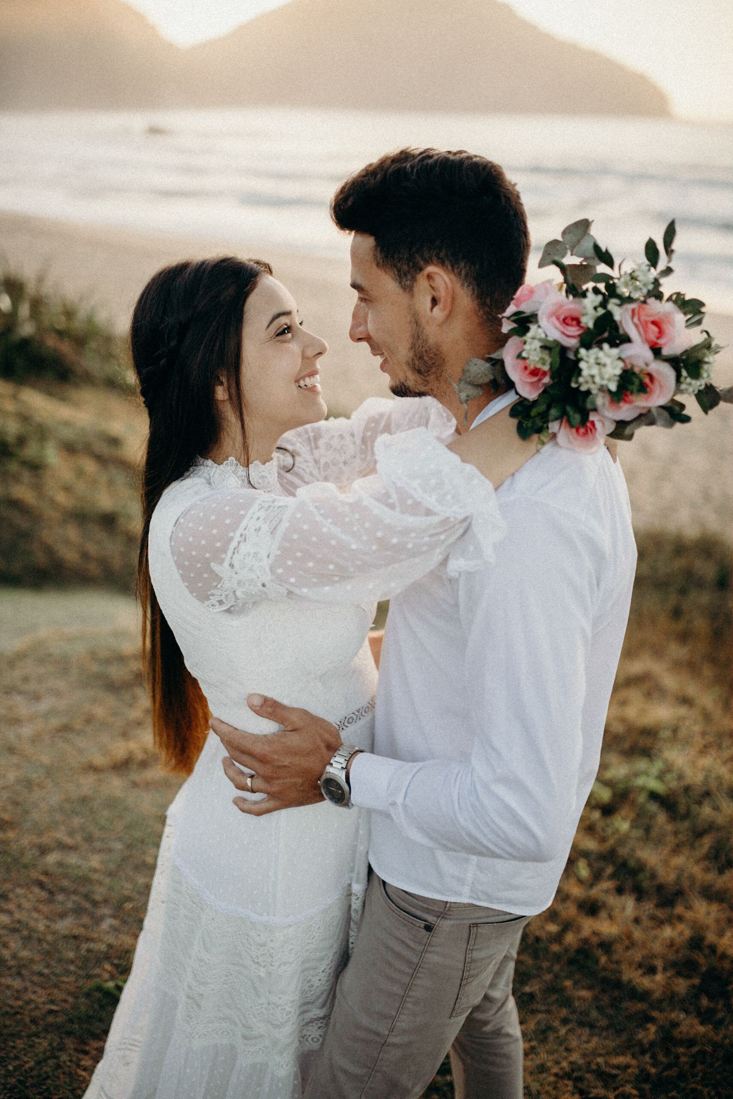 Couple in white and grey embracing near floral arrangement on beach.