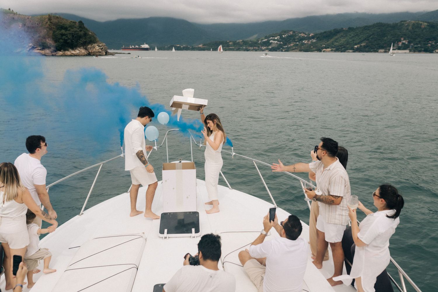 A group celebrates on the bow of a yacht with blue smoke effects against a mountainous coastal backdrop.