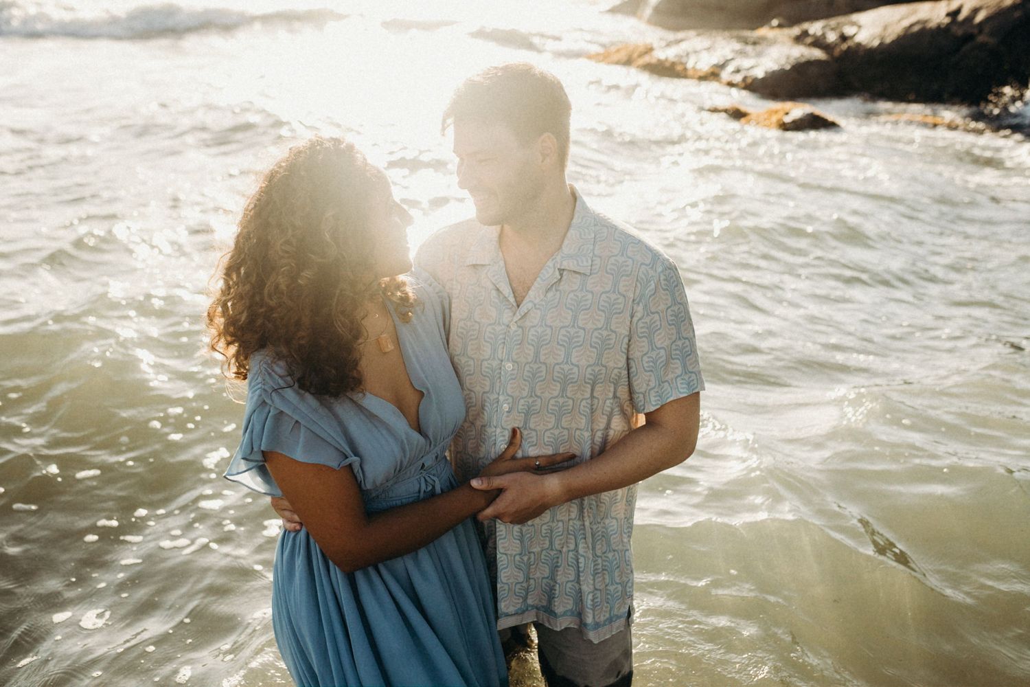 Couple embracing on beach with sunlit waves and coastal scenery.