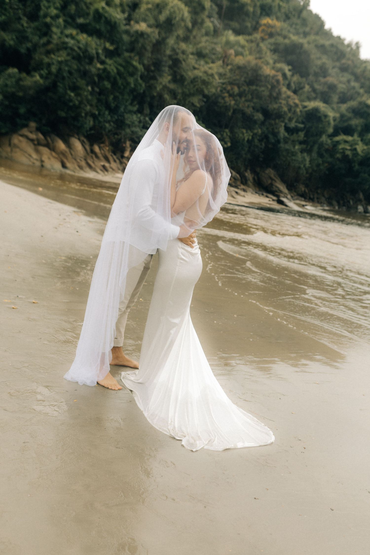 Couple in wedding attire standing in shallow water with flowing veil catching the breeze.