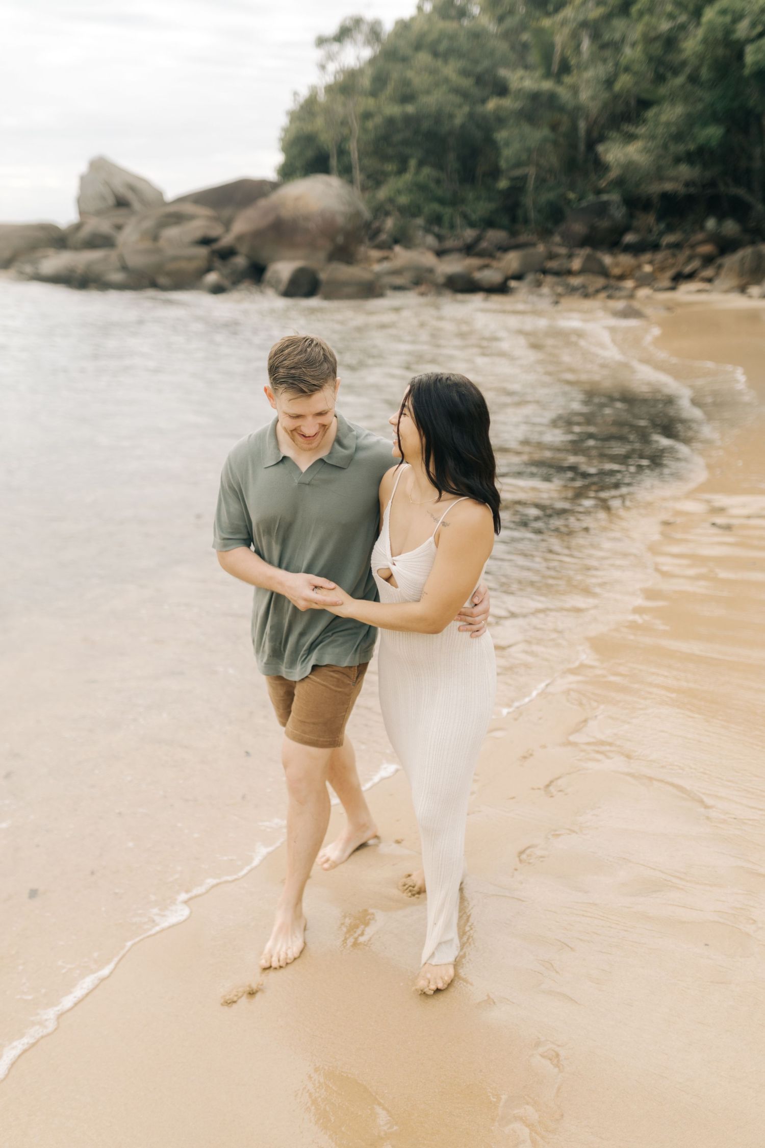A couple walks hand in hand along the shoreline of a secluded beach with rocky formations.