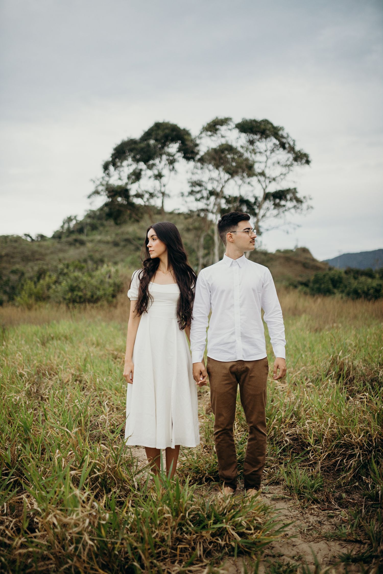 A couple standing in a grassy field with eucalyptus trees and mountains in the background.