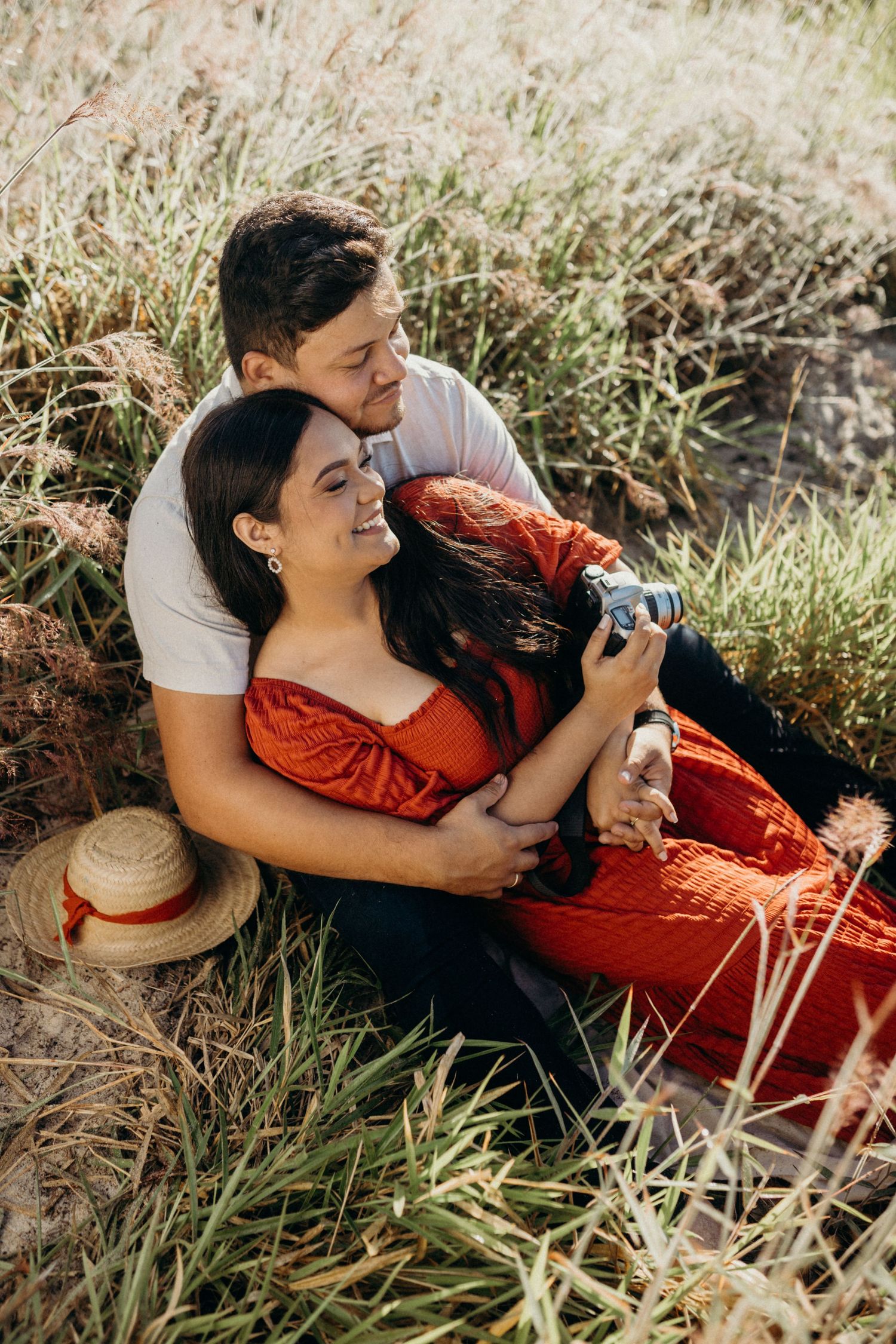 A tender moment between a couple sharing a camera while sitting in tall grass at sunset.