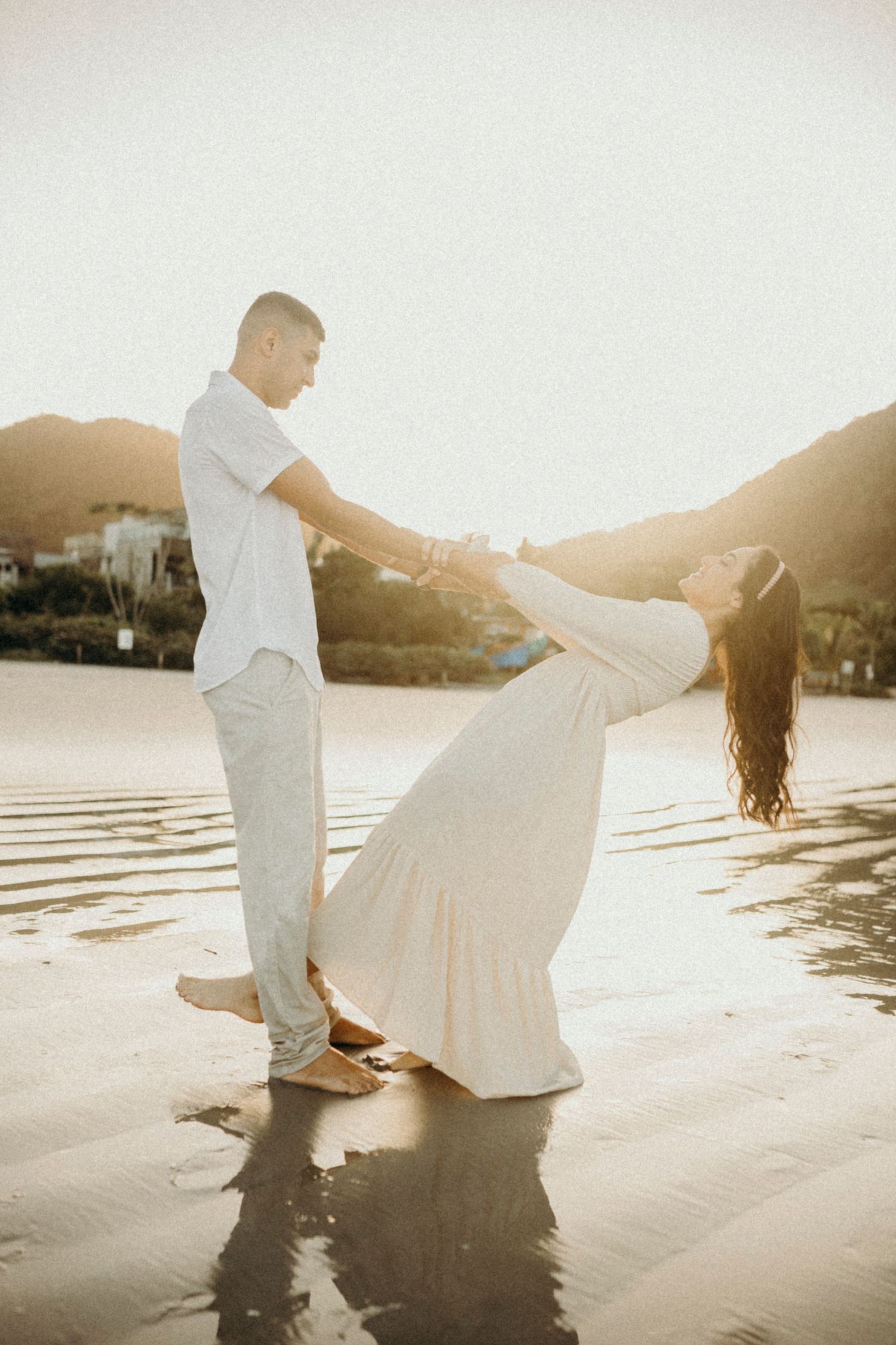 A couple in white outfits dancing playfully on a beach at sunset.