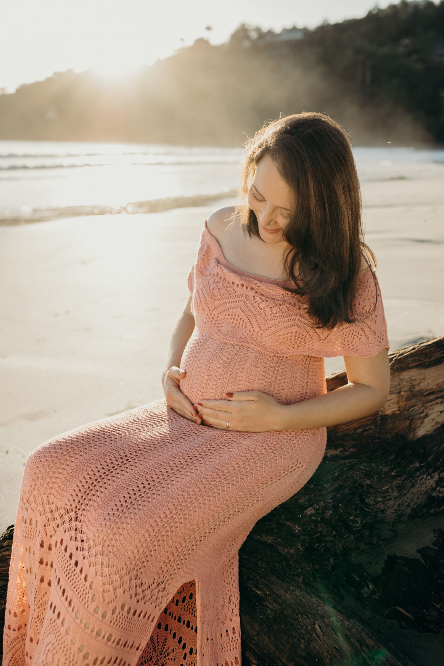 Woman in coral crochet dress sitting on coastal rocks during sunset.
