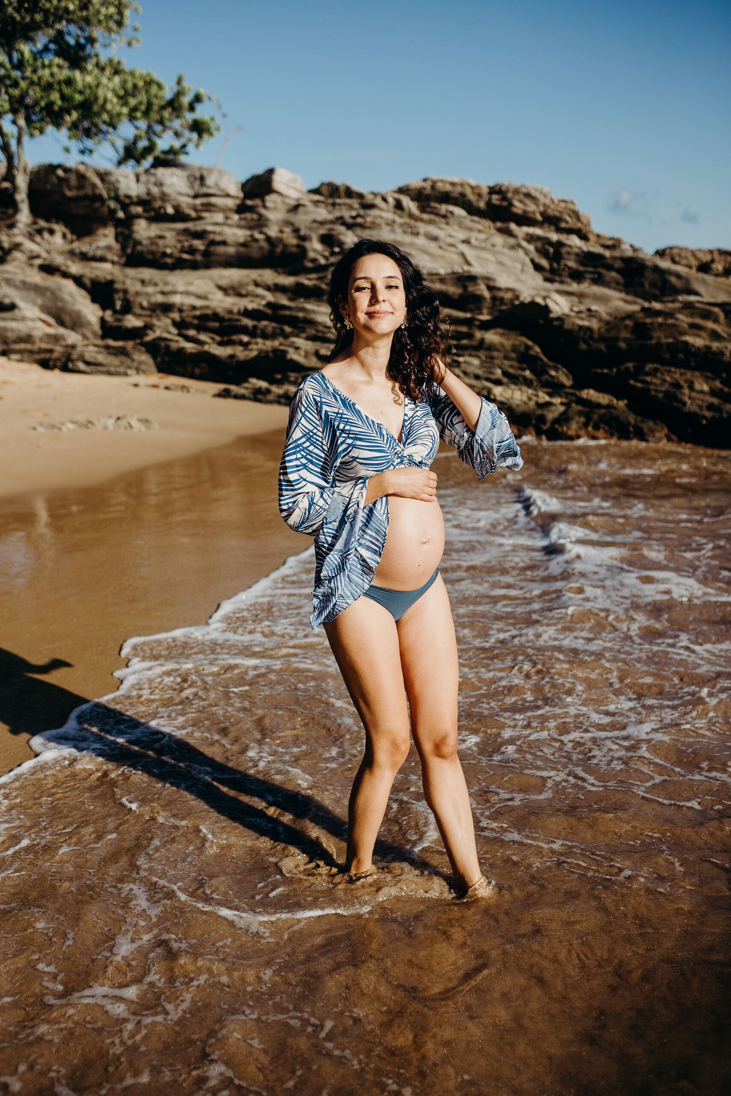A serene maternity photo with crashing waves and volcanic rocks on a tropical beach.