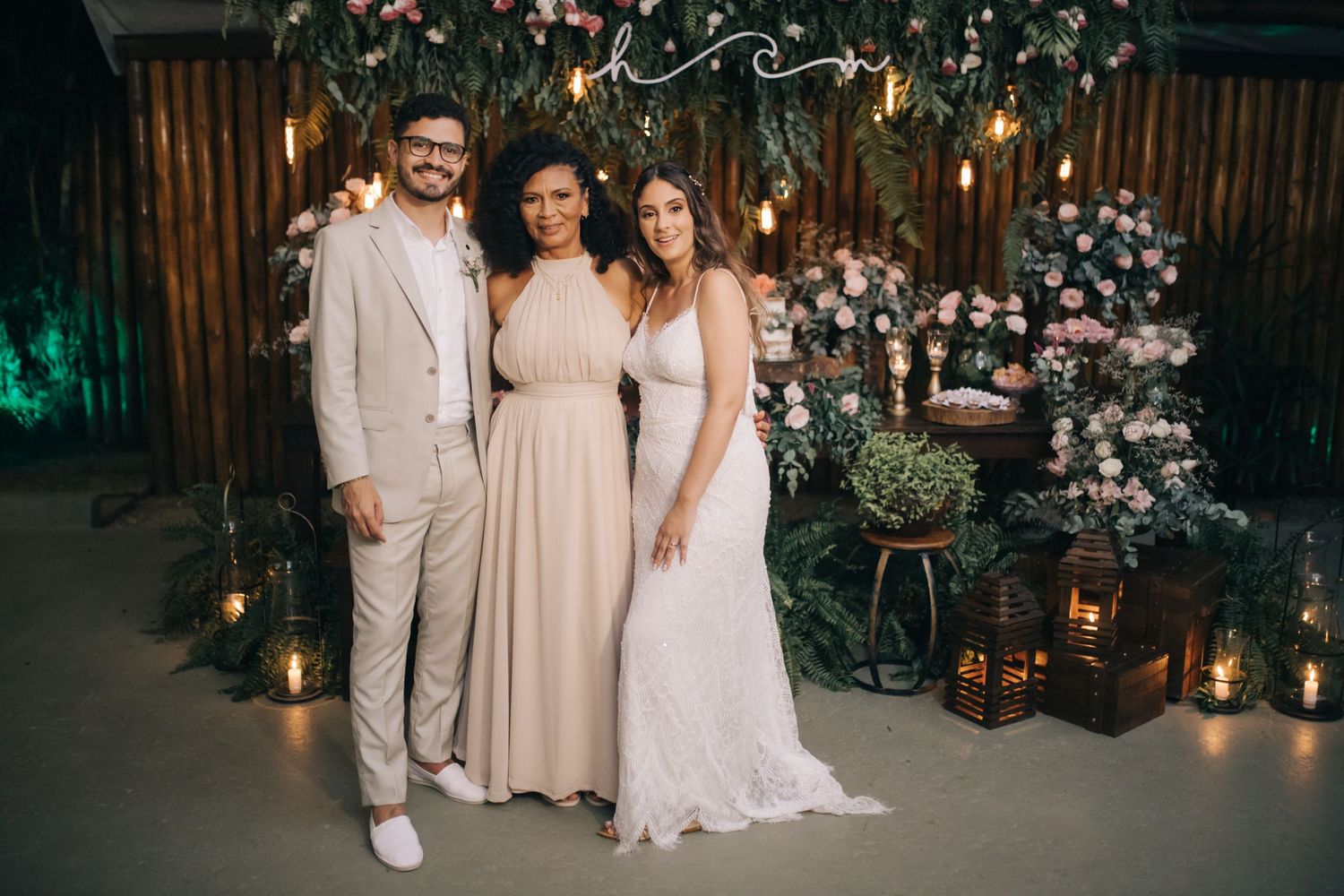 Wedding party group poses in front of floral wall with romantic lighting and lanterns at indoor venue.