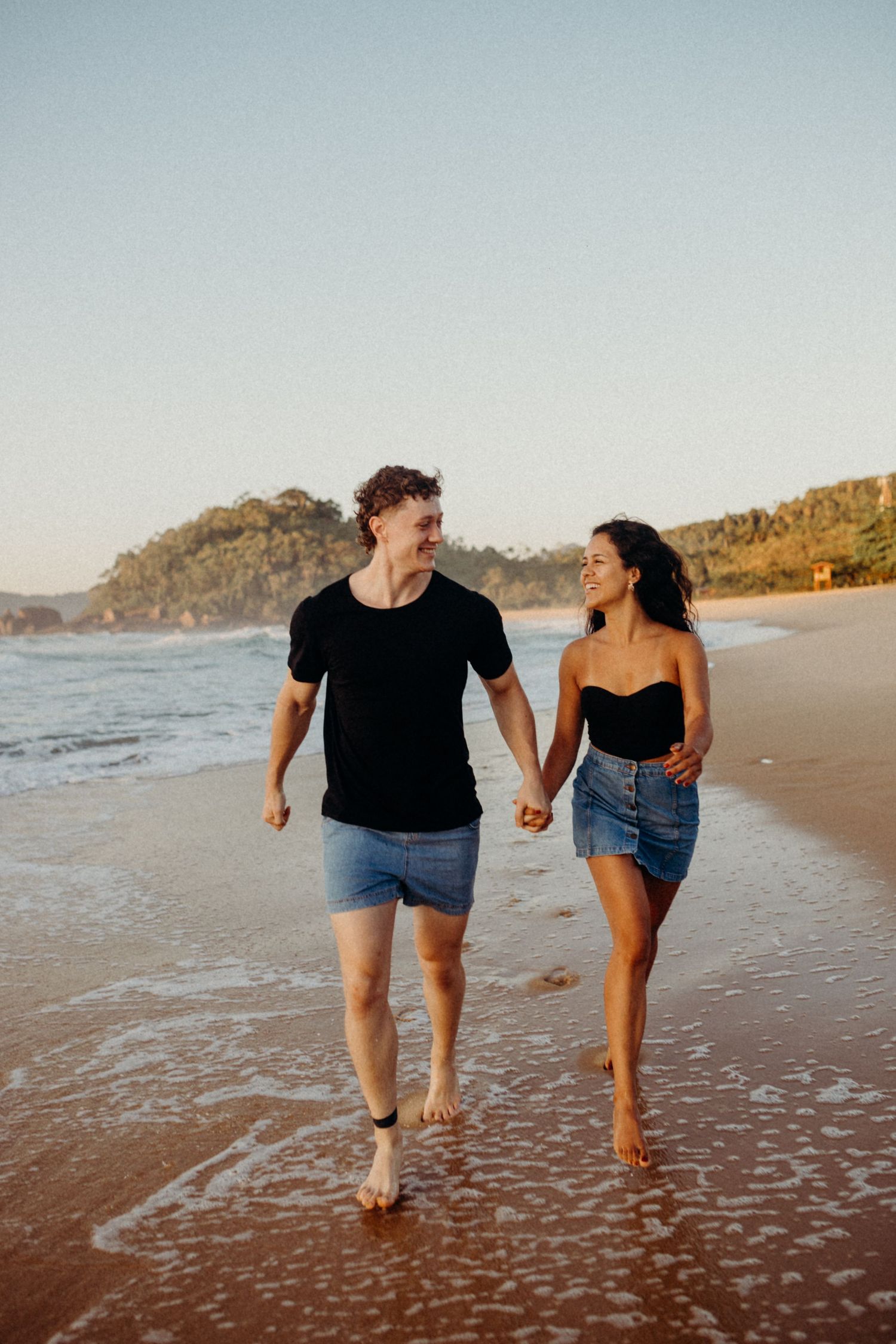 Couple walking hand in hand along the shoreline during golden hour at a scenic beach.