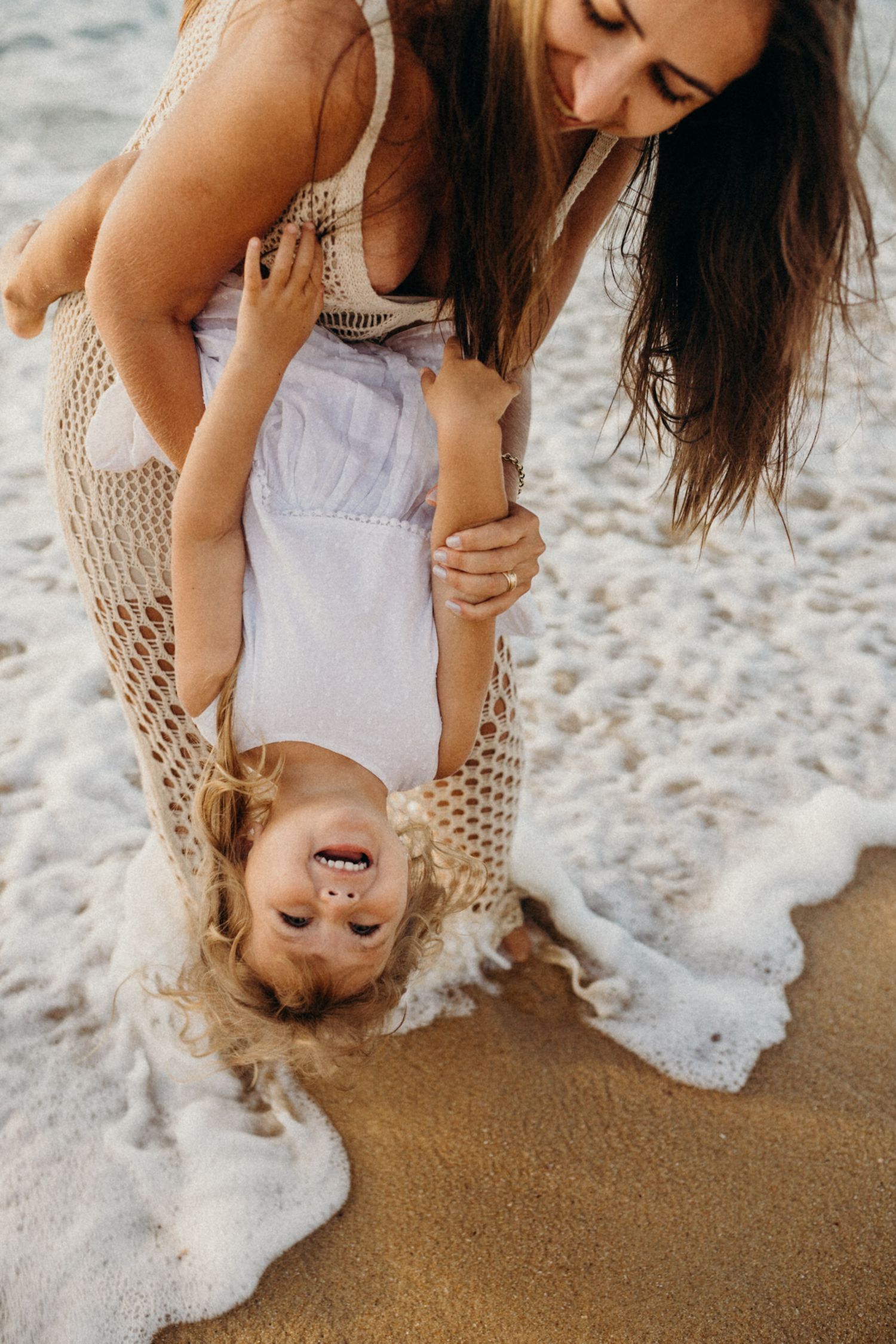 Joyful beach playtime with a child held upside down near ocean waves.