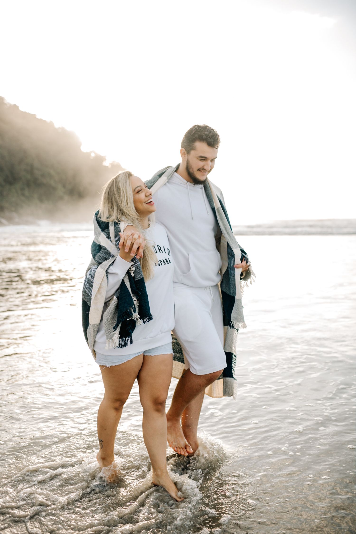 A couple wades through shallow ocean waves while wearing casual beach attire.