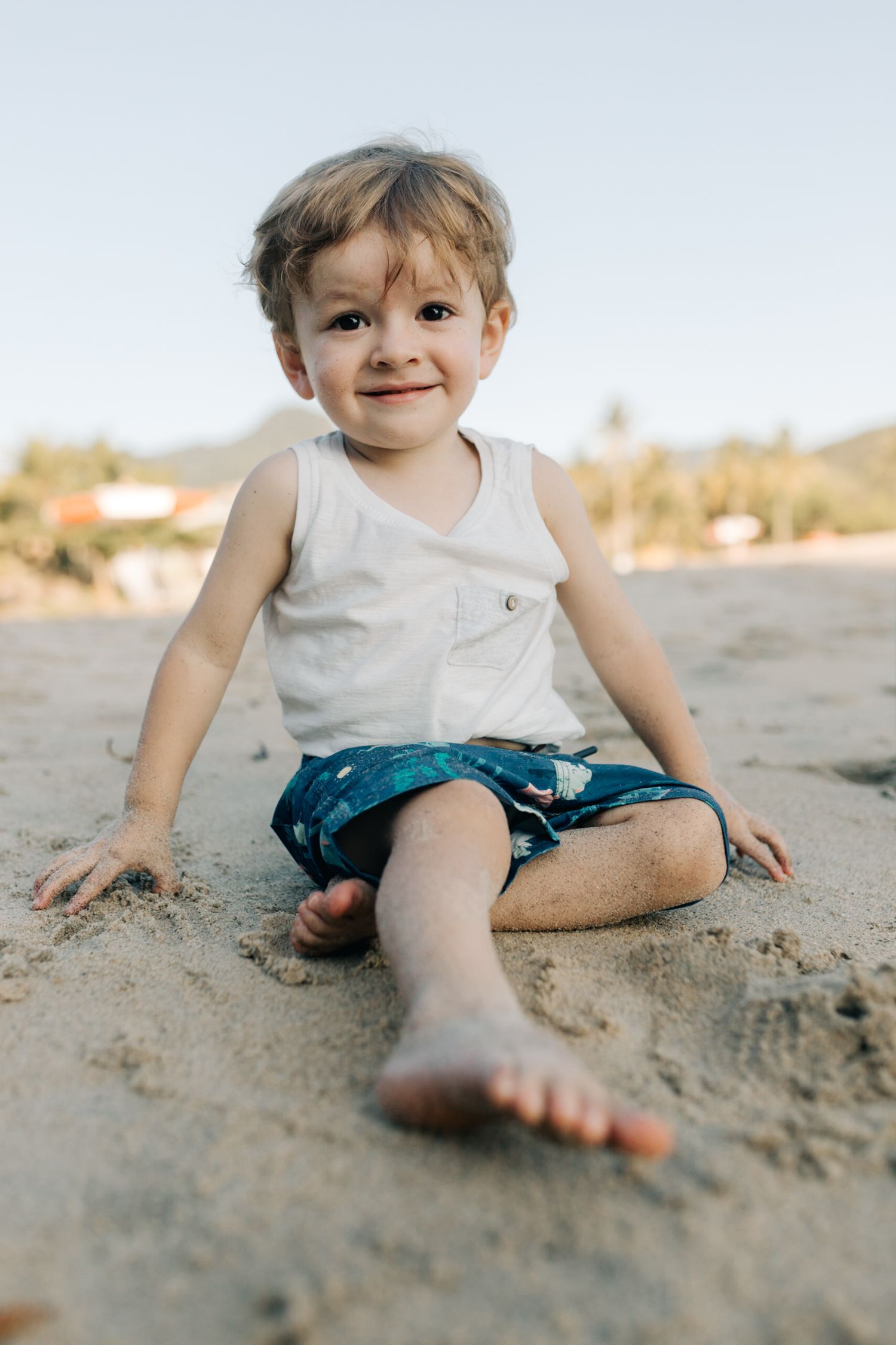 Child sitting on sandy beach wearing blue shorts and white tank top with mountains in background.