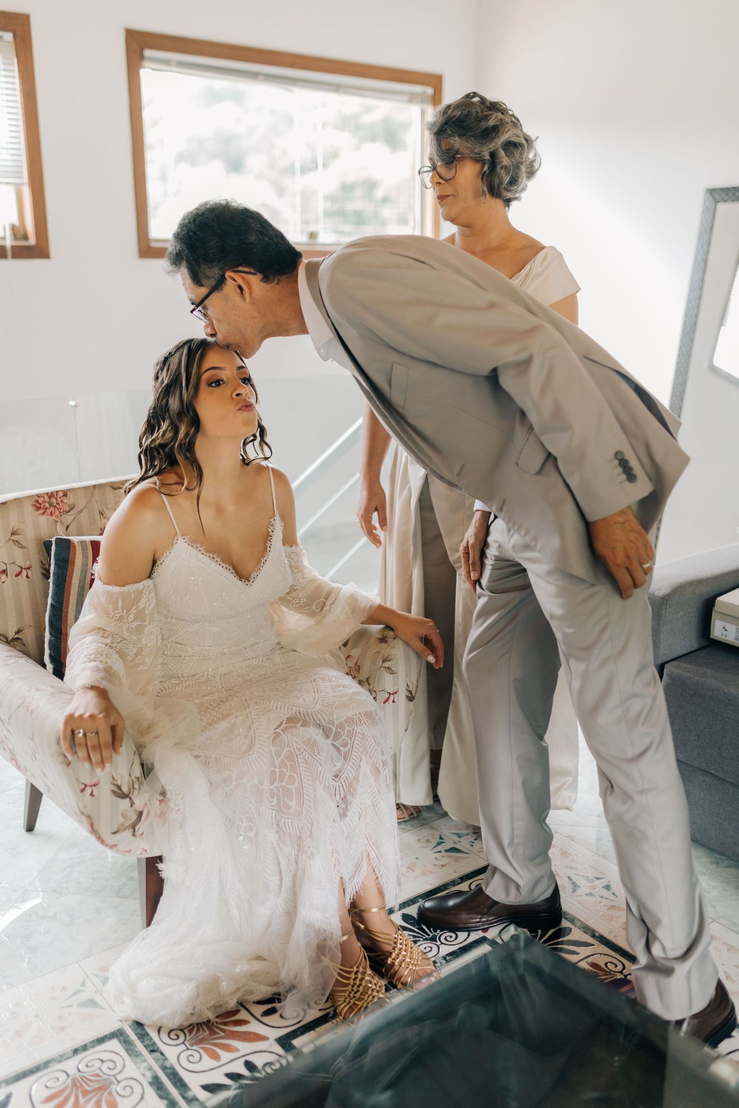 A bride in a lace wedding dress sits while getting ready with help adjusting her dress.