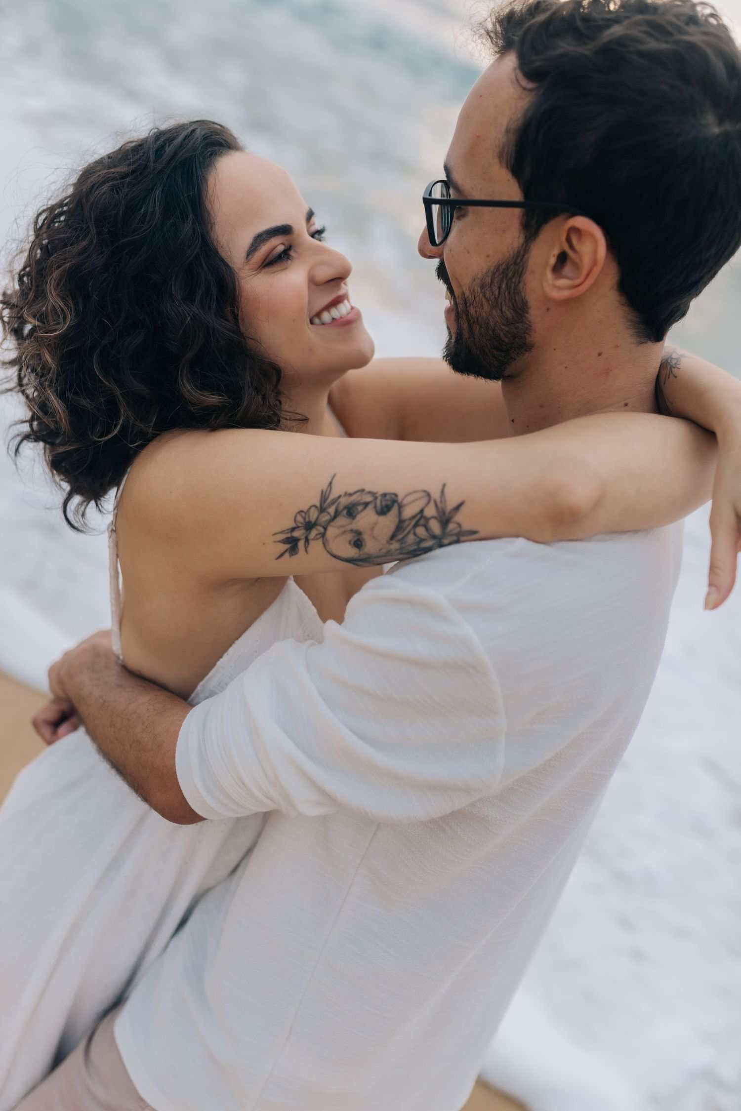 A joyful couple embraces on the beach, dressed in white against the ocean backdrop.