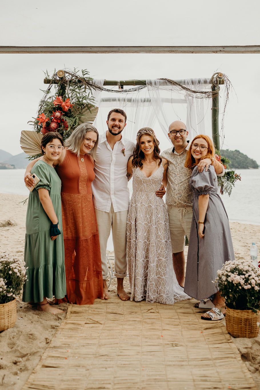 Wedding guests gather under a floral arch on a beach boardwalk at golden hour.