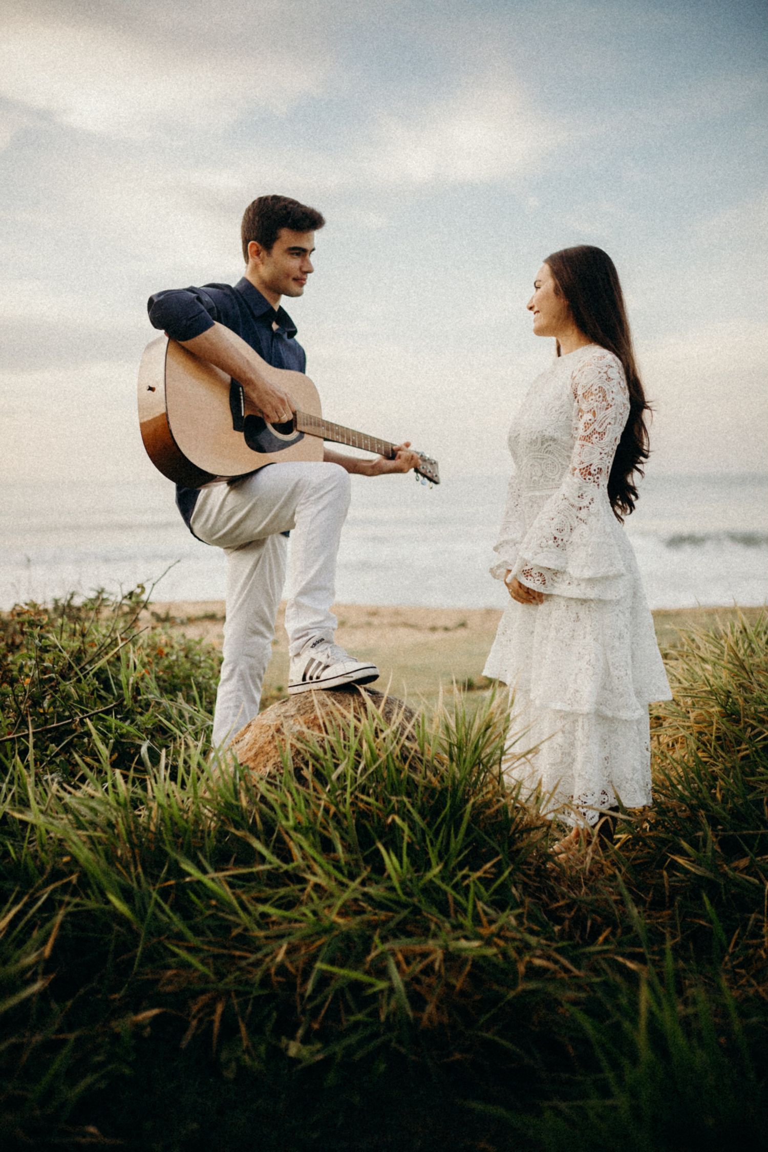 A couple in navy and white lace share a musical moment on a coastal hillside.