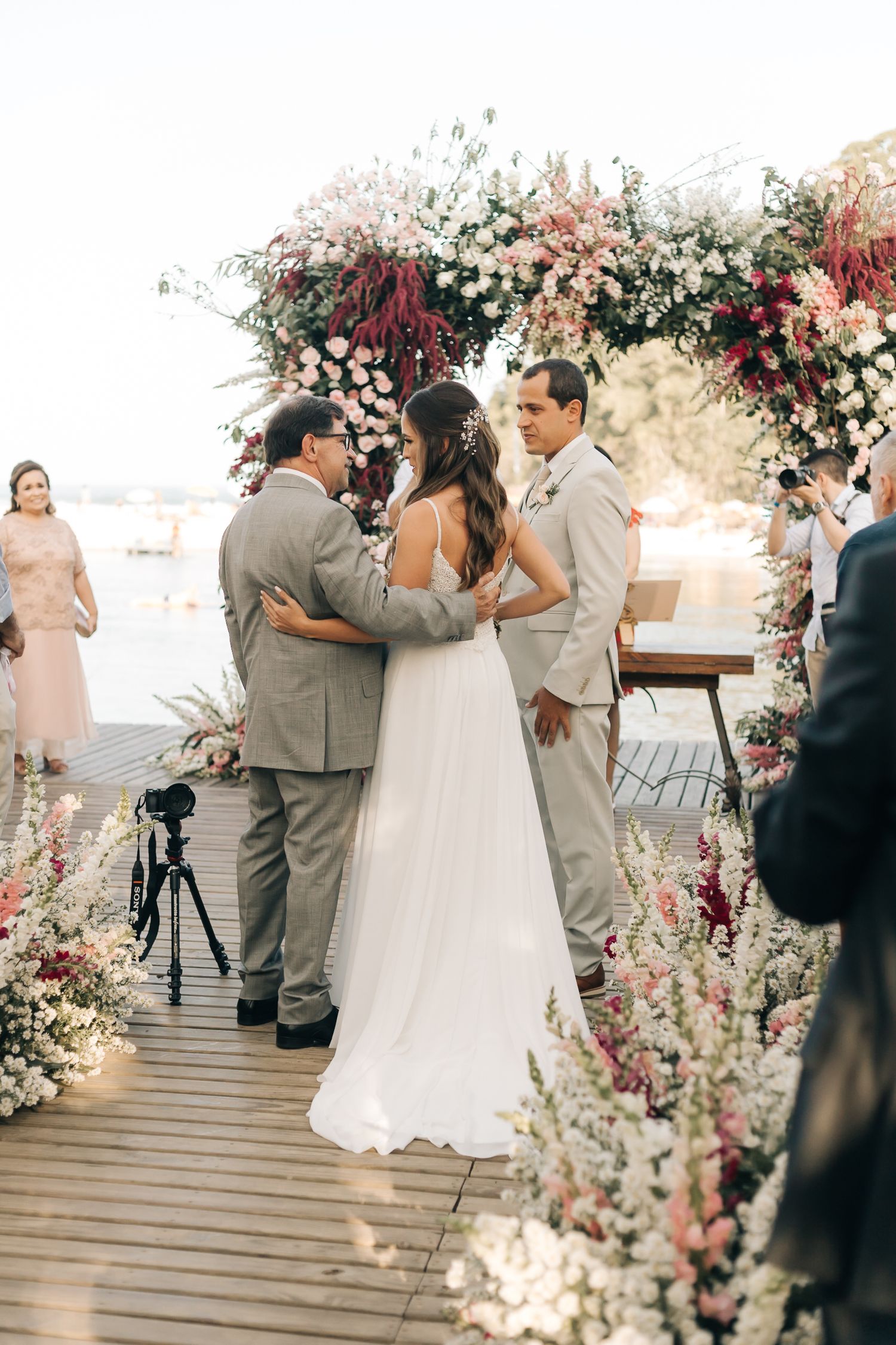 Wedding ceremony under floral arch on lakeside wooden deck with pink and white blooms.