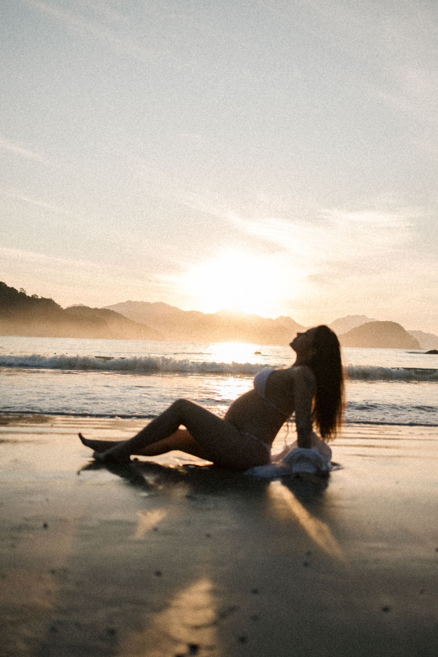 Silhouette of an expectant mother posing on the beach at sunset with mountains in the background.