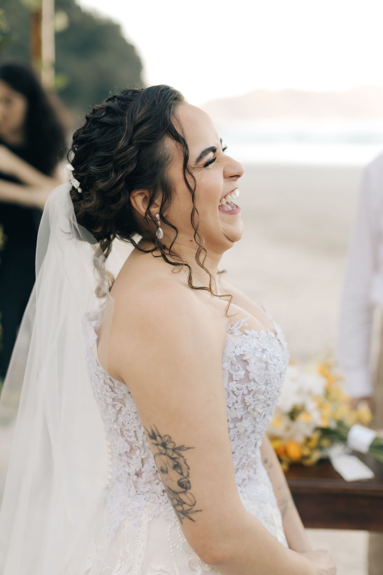 A bride in a lace wedding dress laughs joyfully at her beach wedding ceremony.