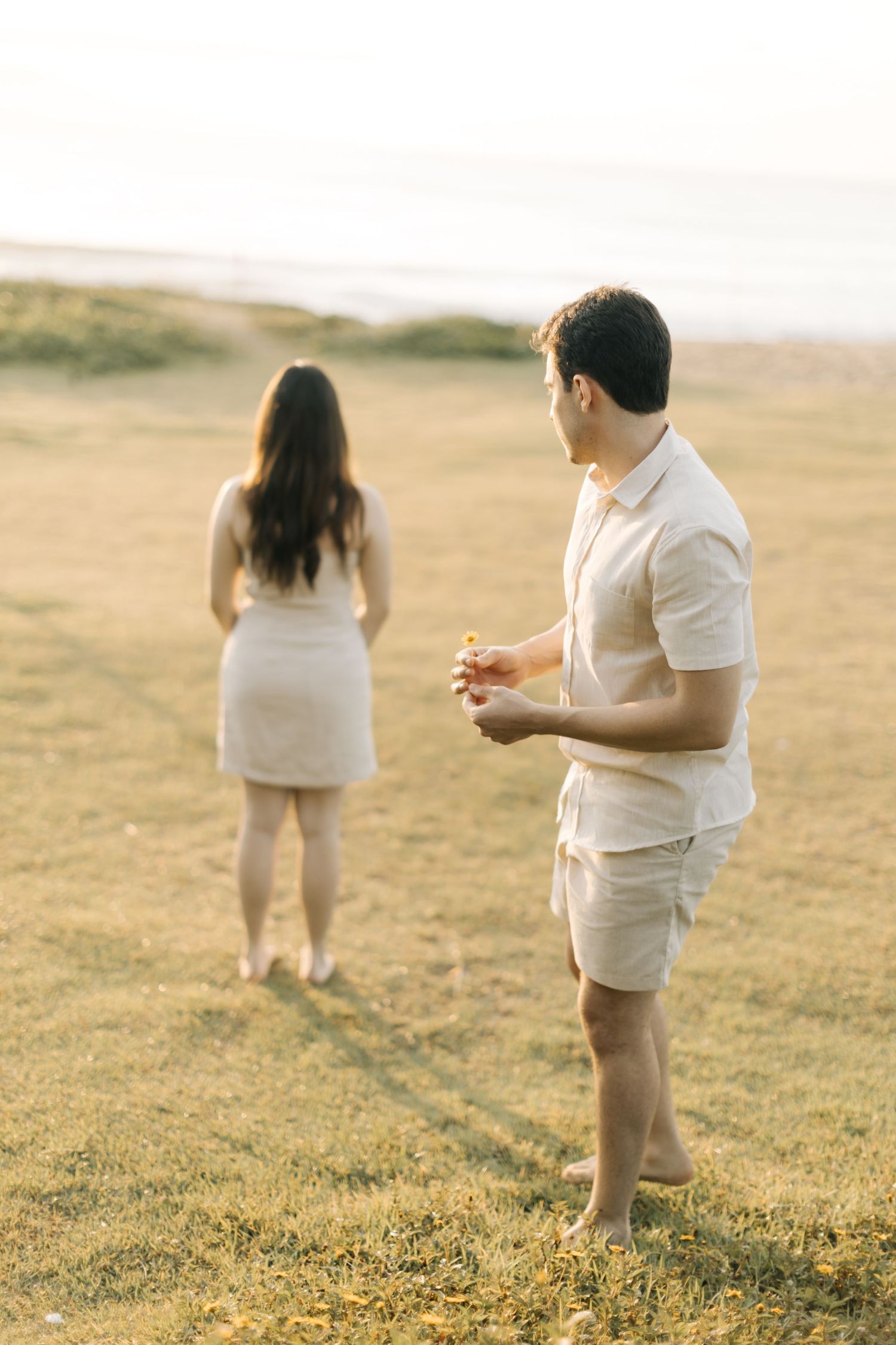 Couple in light colored outfits stands in a golden field during a surprise proposal moment.