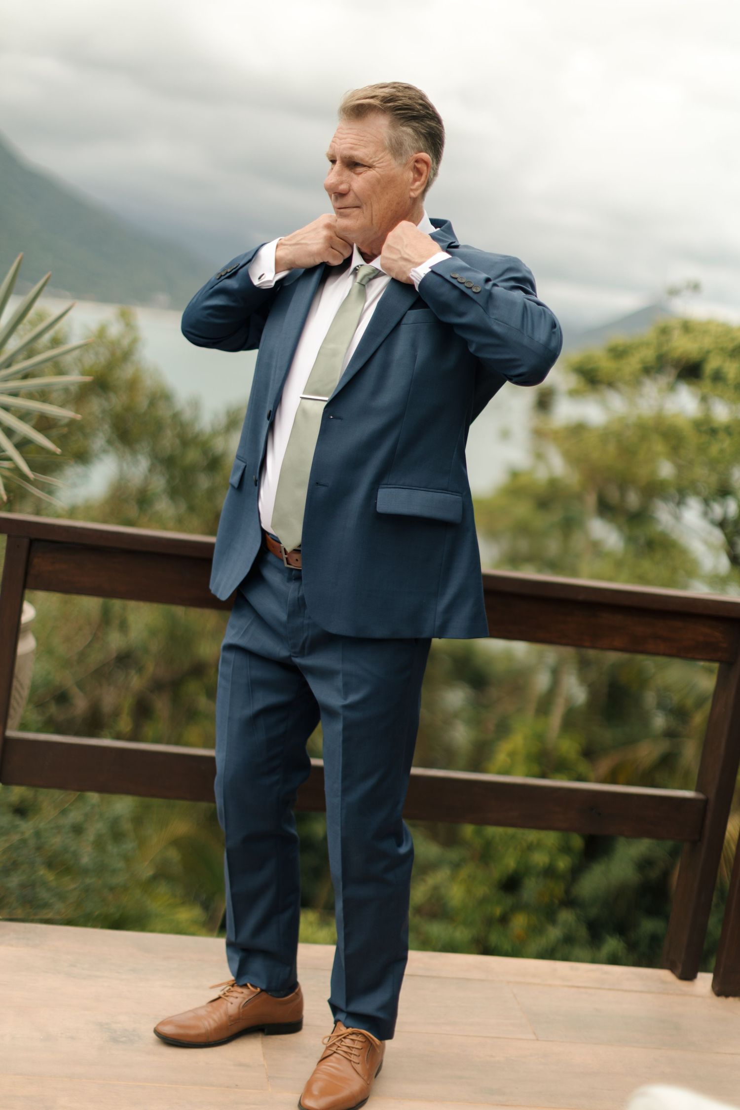 A person in a navy blue suit and brown shoes poses playfully on an outdoor deck with mountain views.