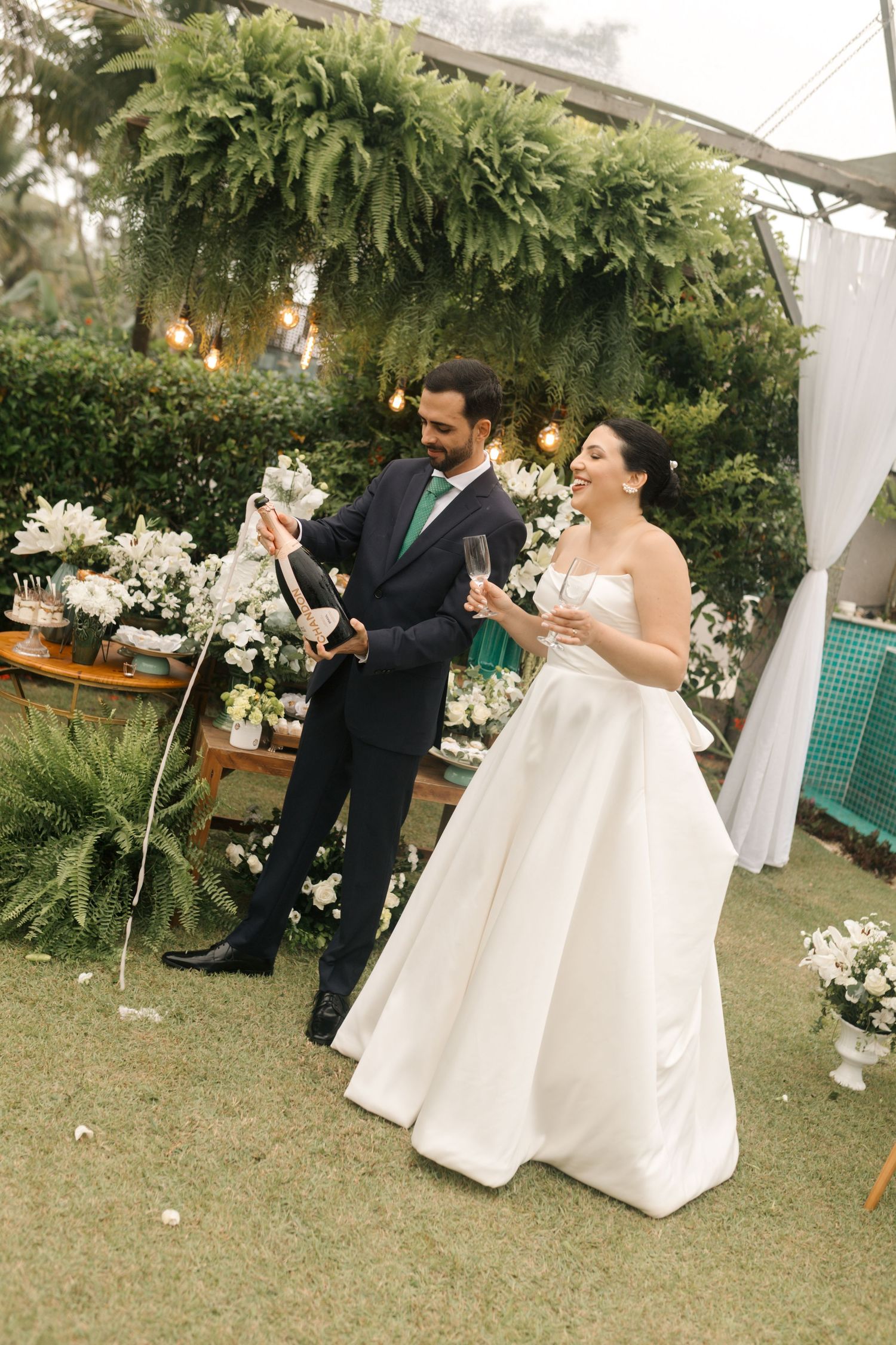 Wedding couple toasting under a greenery-covered arch with white floral decor.