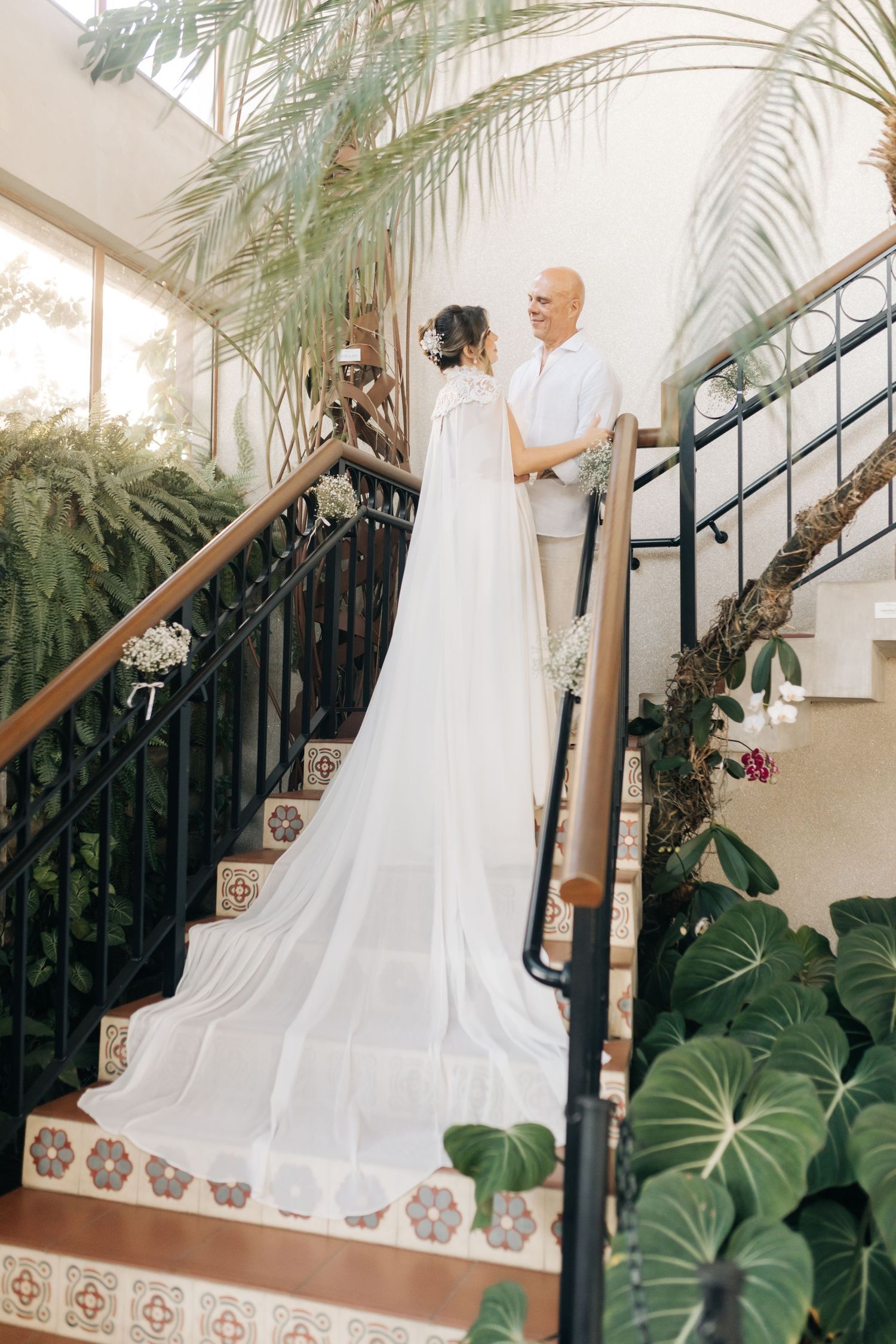 A couple poses on an elegant staircase decorated with tropical plants and palm fronds.