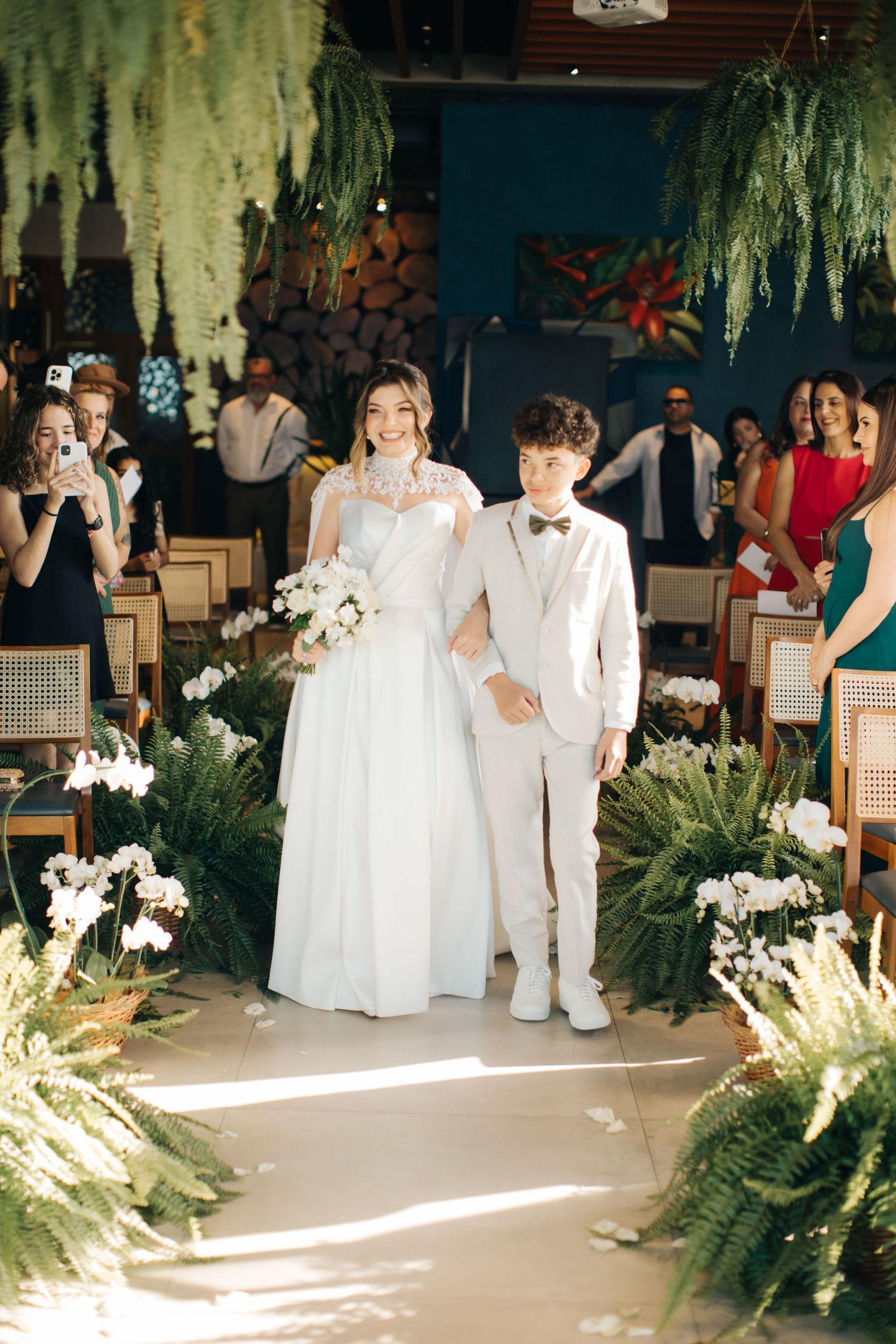 Wedding ceremony setup with hanging ferns, white flowers and wooden chairs in an elegant indoor venue.