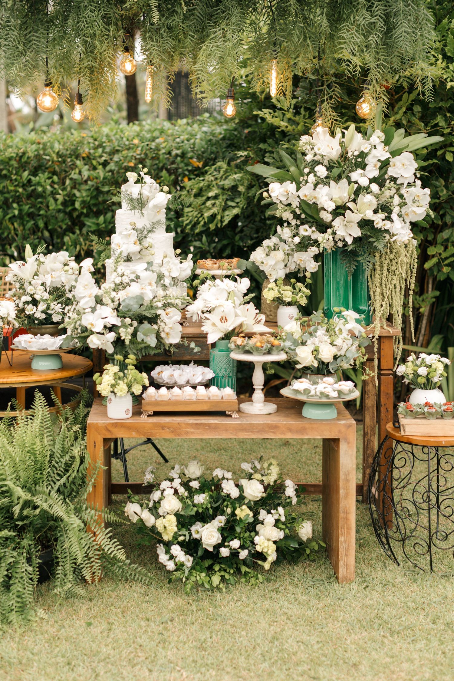 Rustic outdoor dessert display with wooden tables adorned with white floral arrangements and hanging lights.